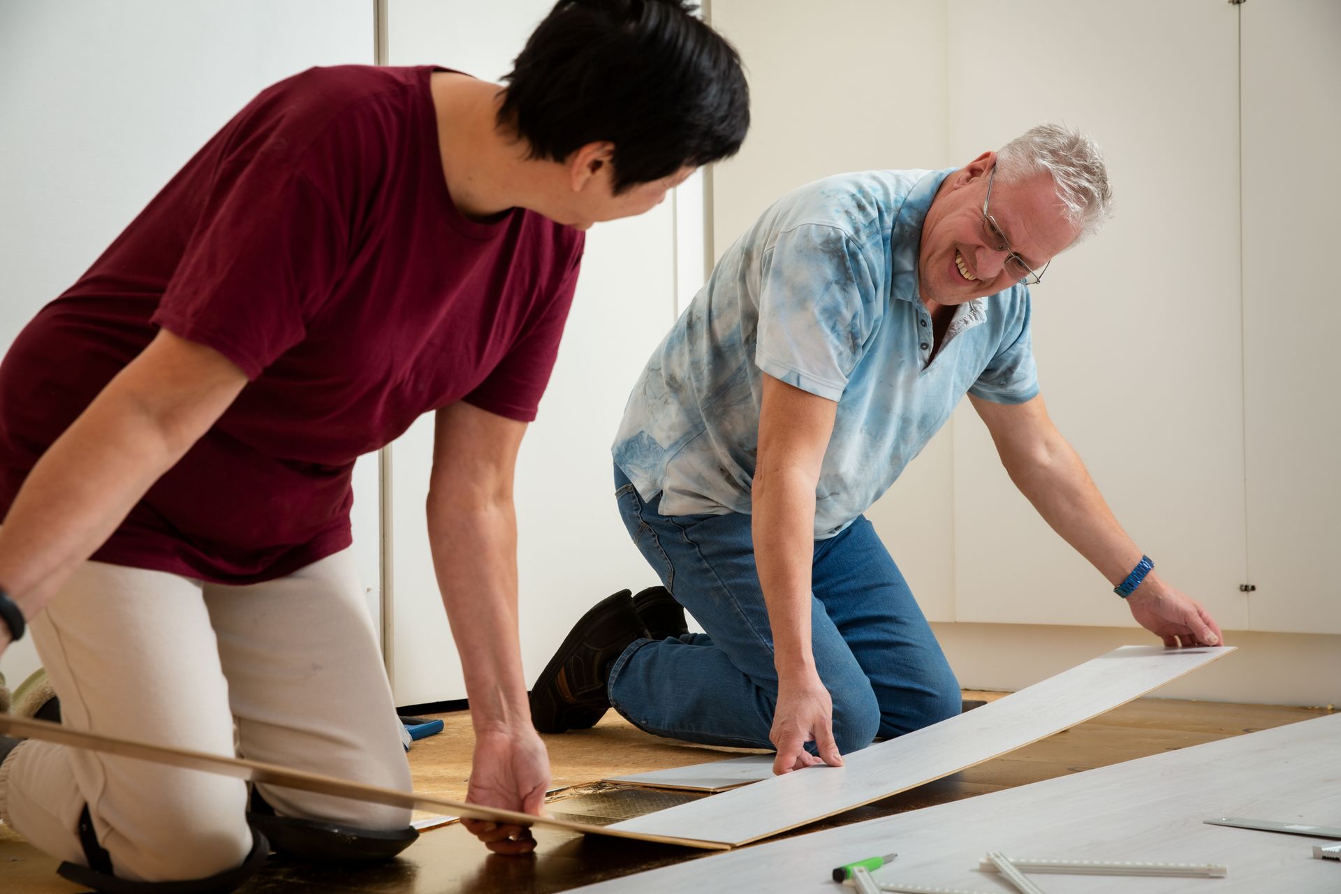 Two people installing flooring, kneeling, looking at a plank. One wears a maroon shirt, the other a blue shirt.