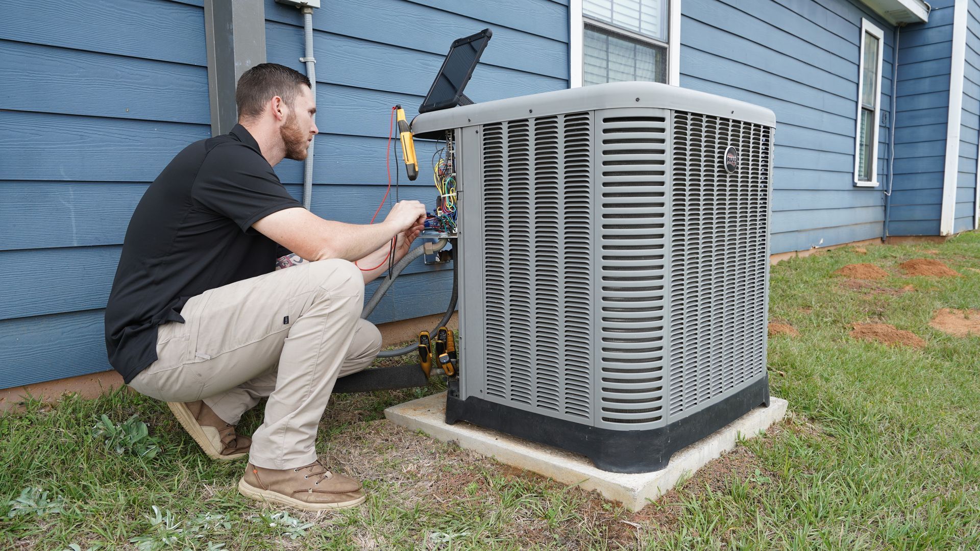 A technician crouching outside next to a residential HVAC unit, inspecting the wiring with a multimeter.