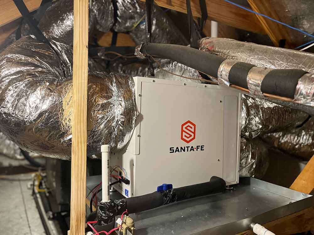 A white Santa Fe brand dehumidifier sits on a metal drip pan in an attic, connected to silver insulated ductwork.