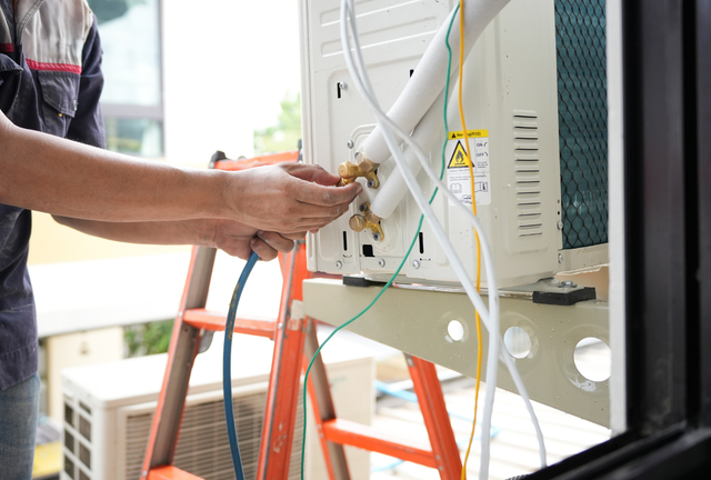 A technician connects a service hose to the exterior unit of an air conditioning system while standing on a ladder.