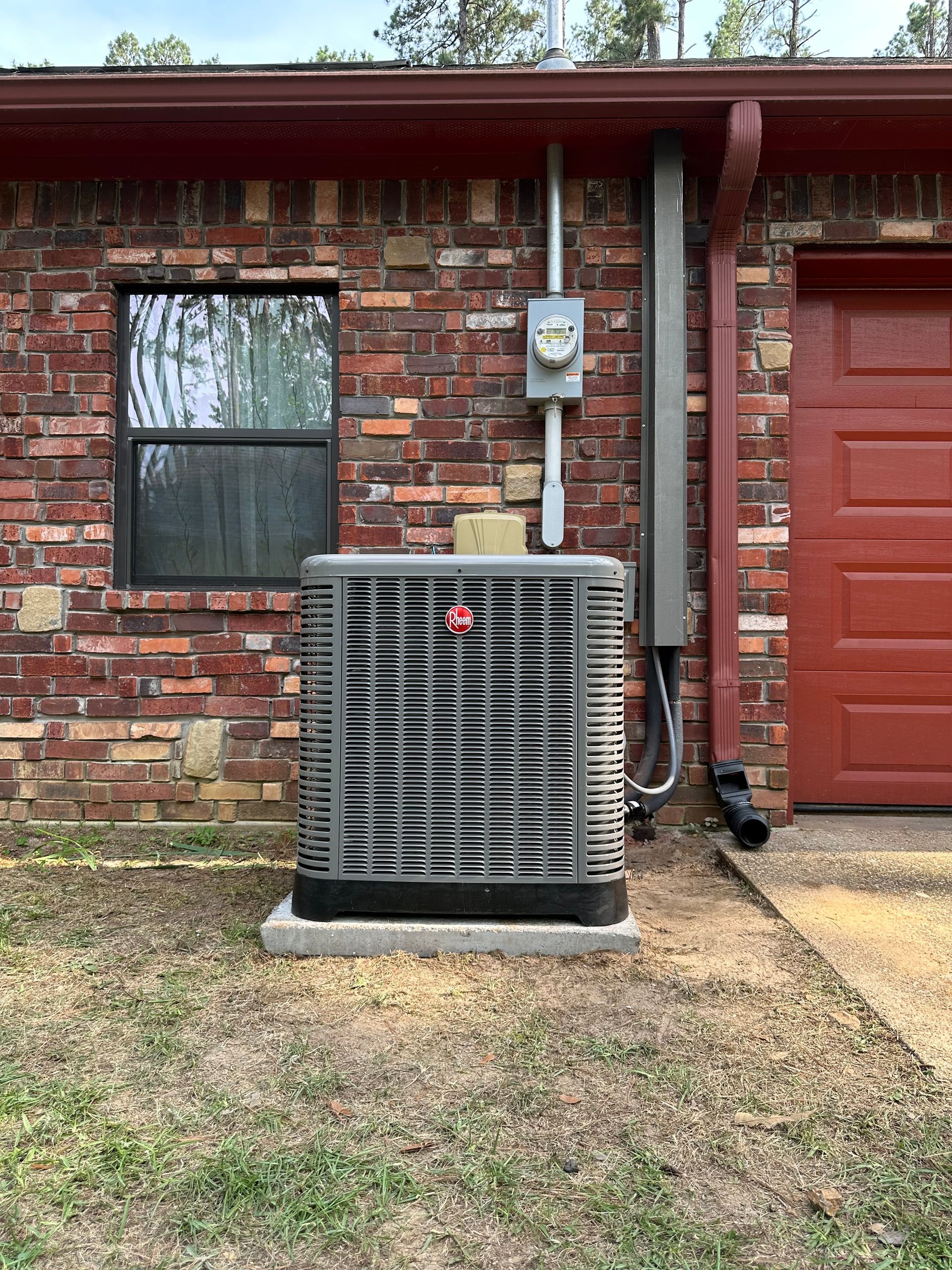 An outdoor air conditioning unit sits on a concrete pad against a brick exterior wall near a door and window.