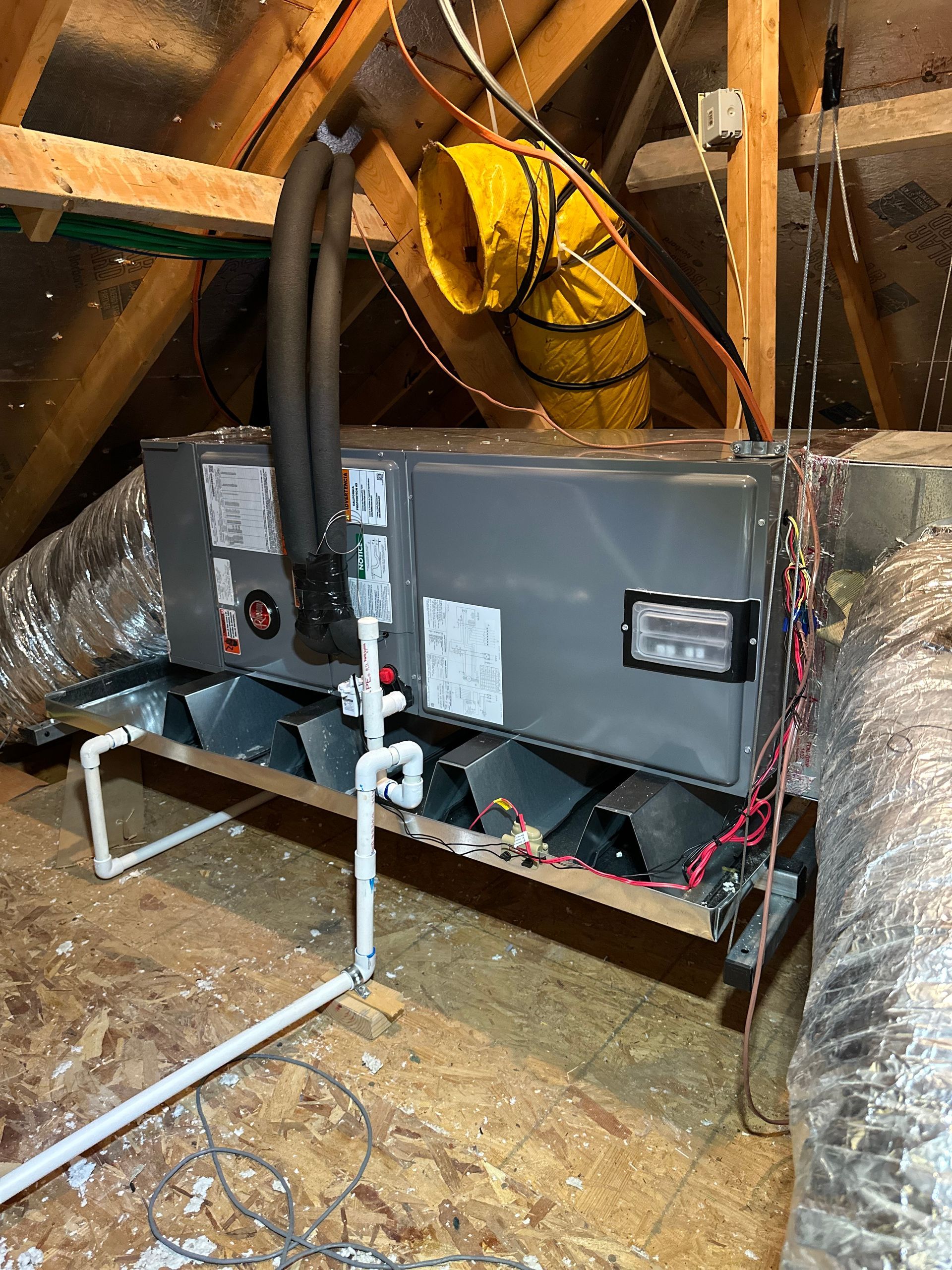 An HVAC air handler unit installed in an attic, featuring connected ductwork, insulated pipes, and white drain piping.
