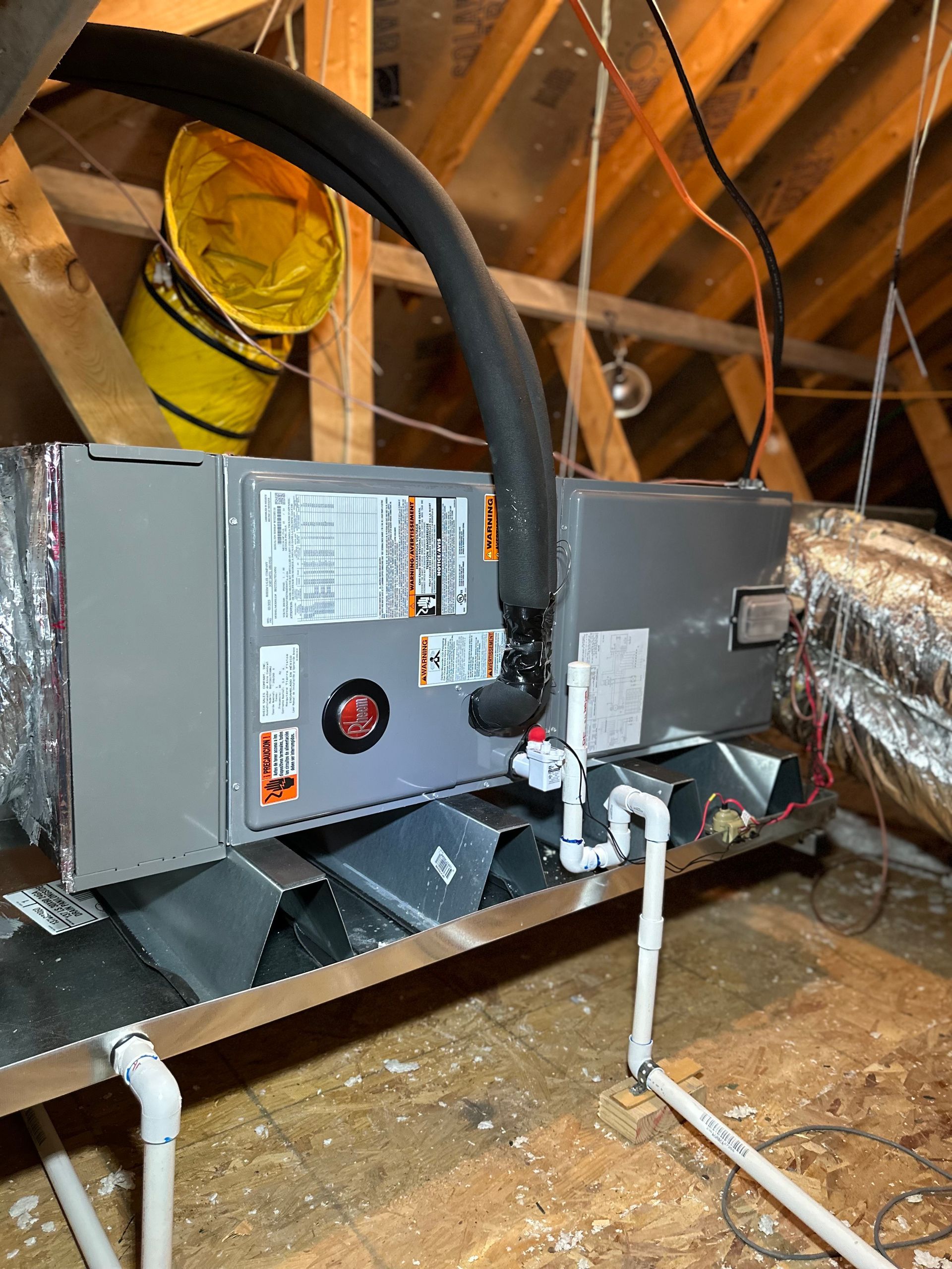 An HVAC air handler unit installed on a metal platform in an attic, featuring connected ductwork and a PVC drain line.