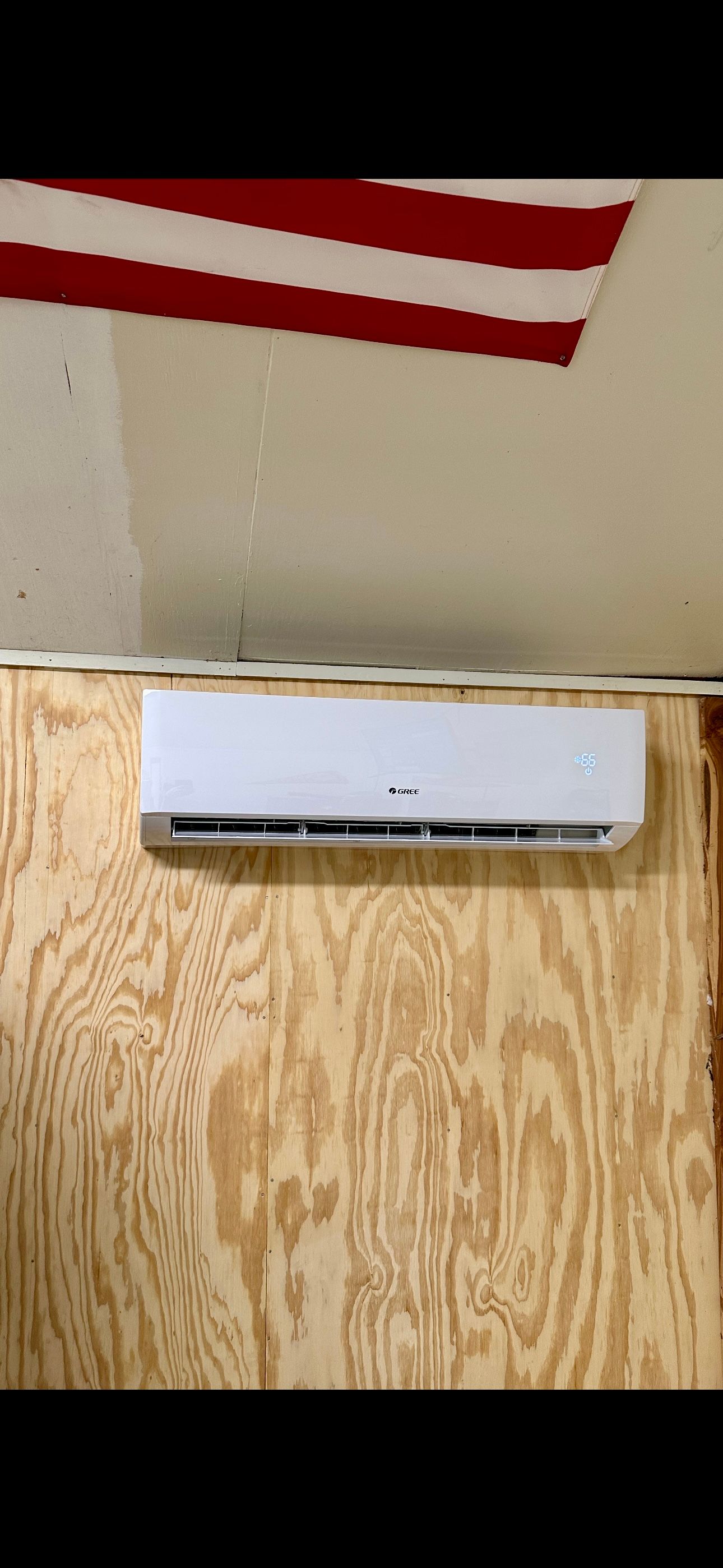 A white mini-split air conditioner mounted on a plywood wall below a partial view of a striped American flag.