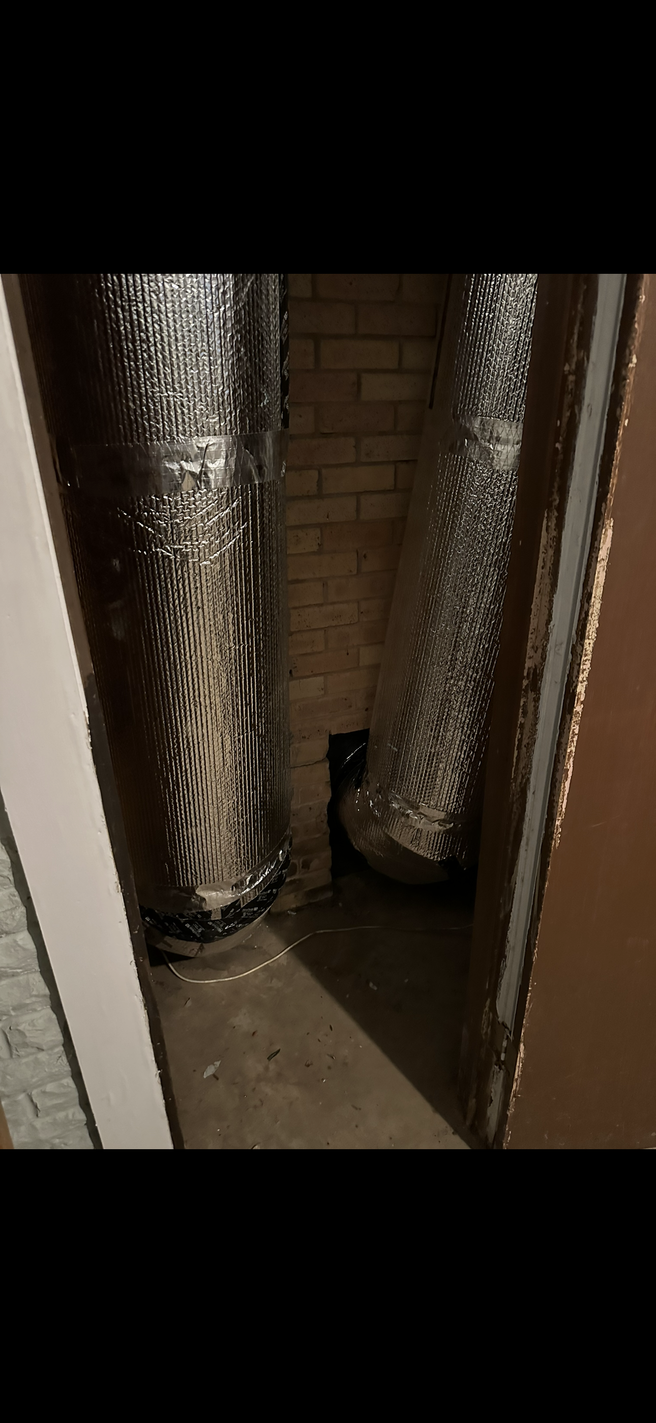 Two metallic insulated air ducts running vertically inside a narrow, unfinished brick utility closet.