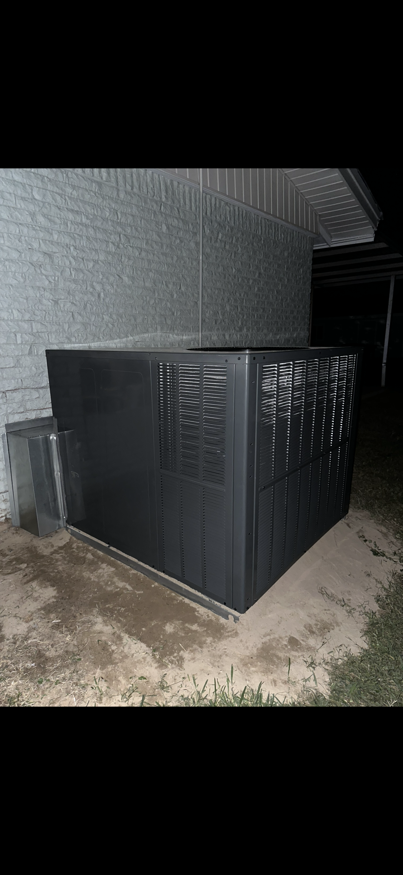 A dark-colored outdoor air conditioning unit sits on a concrete slab next to a grey brick wall at night.