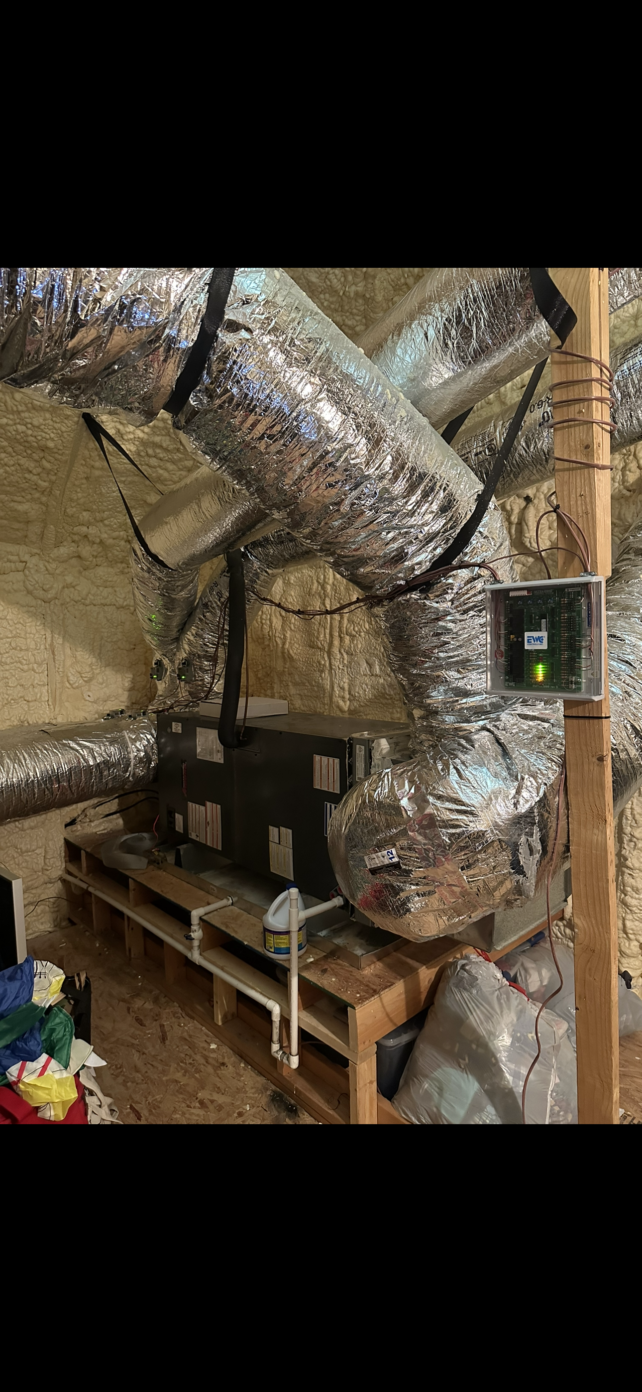 An HVAC air handler and insulated ductwork mounted on a wooden platform in an attic insulated with spray foam.