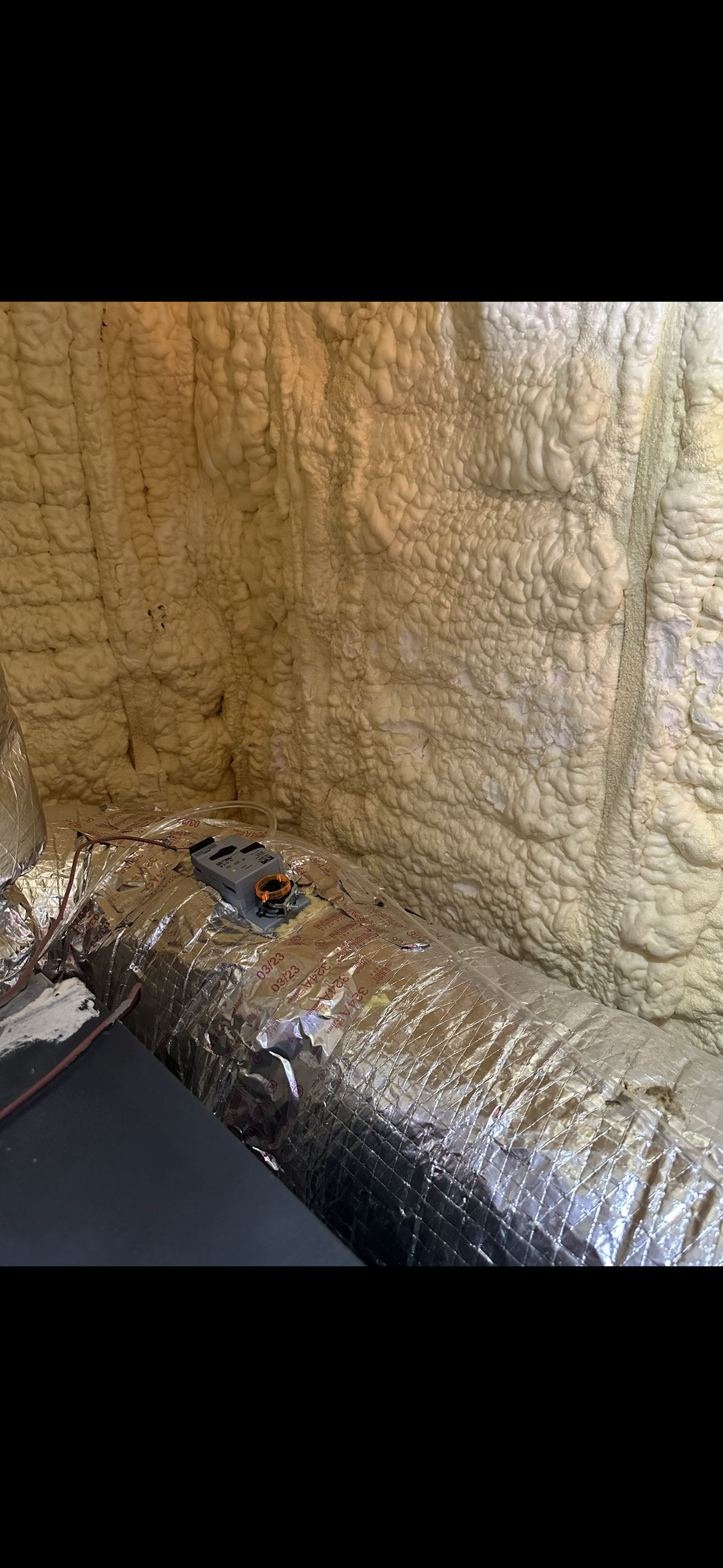 A close-up view of beige spray foam insulation on a wall meeting a silver reflective barrier on the floor.