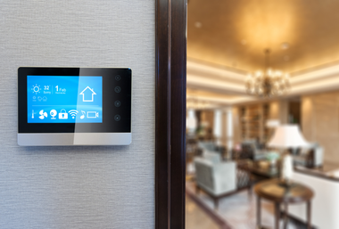 A digital smart home control panel mounted on a textured wall, overlooking a blurred, upscale living room interior.