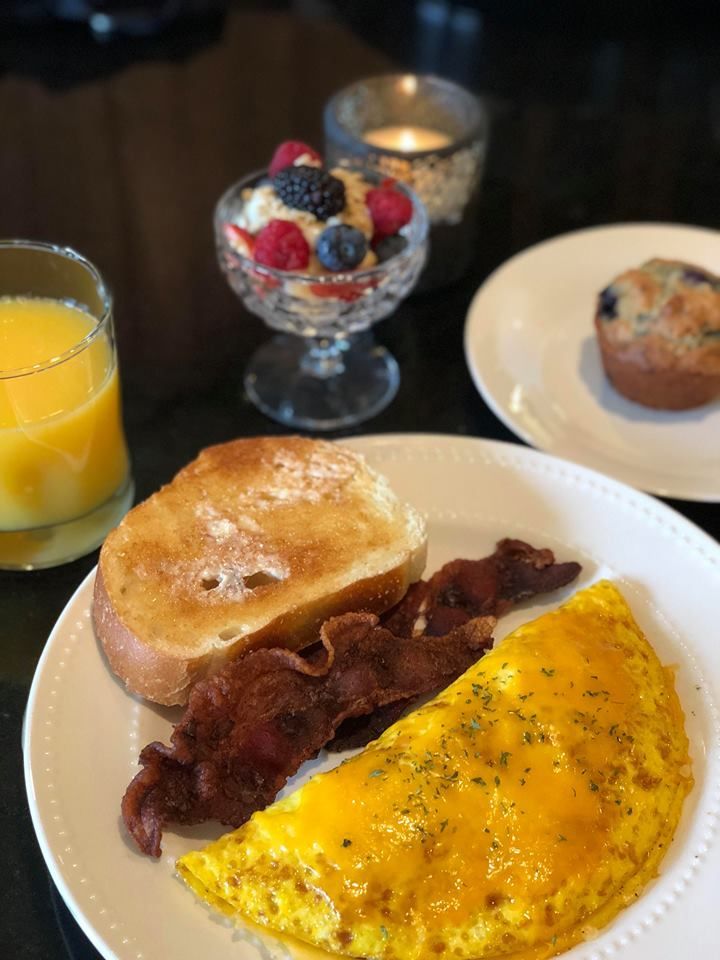A breakfast plate with an omelet, bacon, and toast, alongside orange juice, a fruit cup, and a muffin on a dark table.