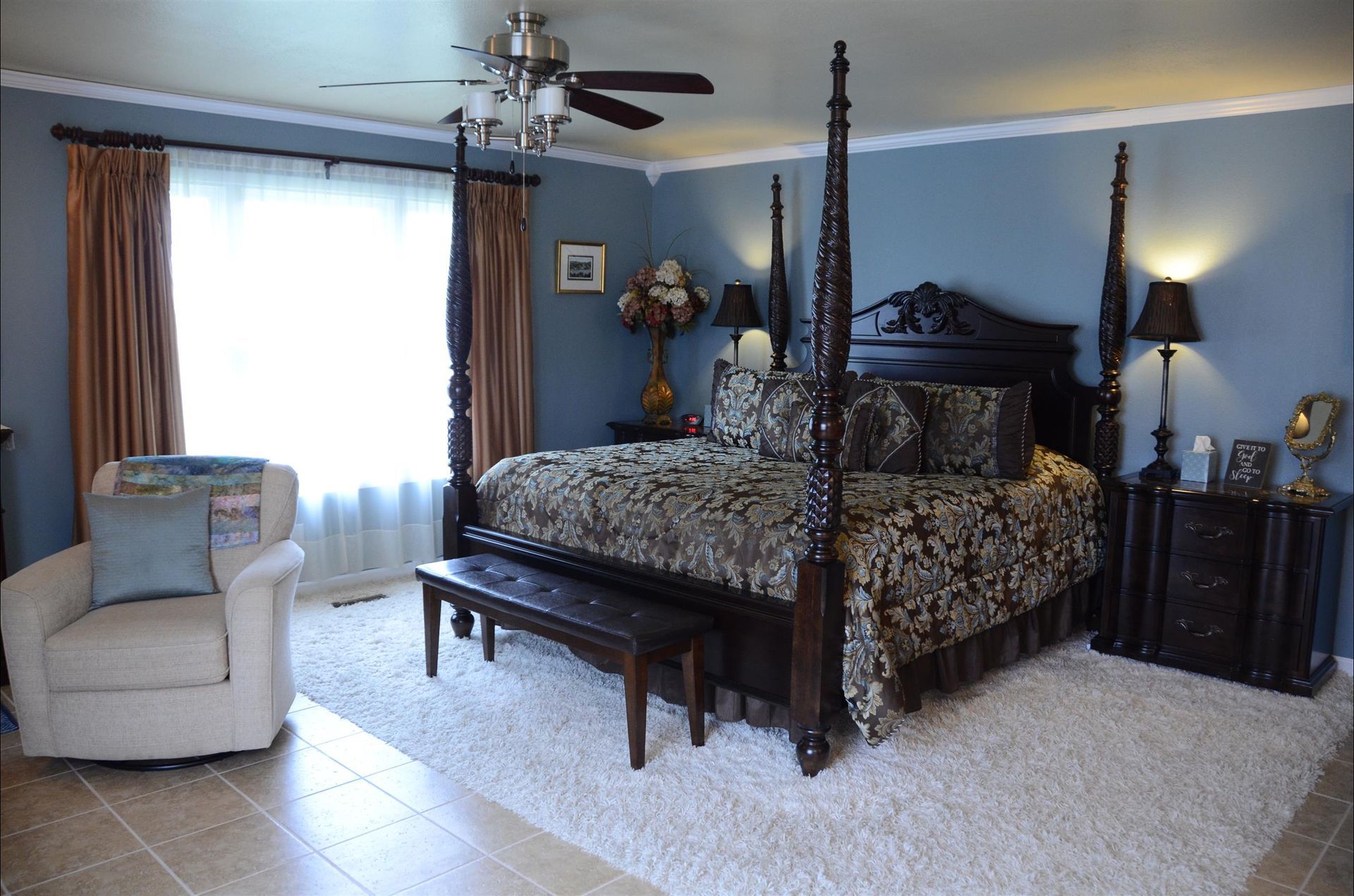 A bedroom with blue walls, a large dark wood four-poster bed, beige carpet, and a light-colored armchair.