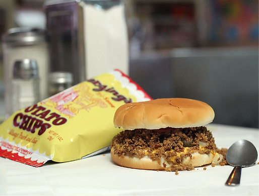 A sloppy joe sandwich on a white surface next to a bag of potato chips and a small spoon.