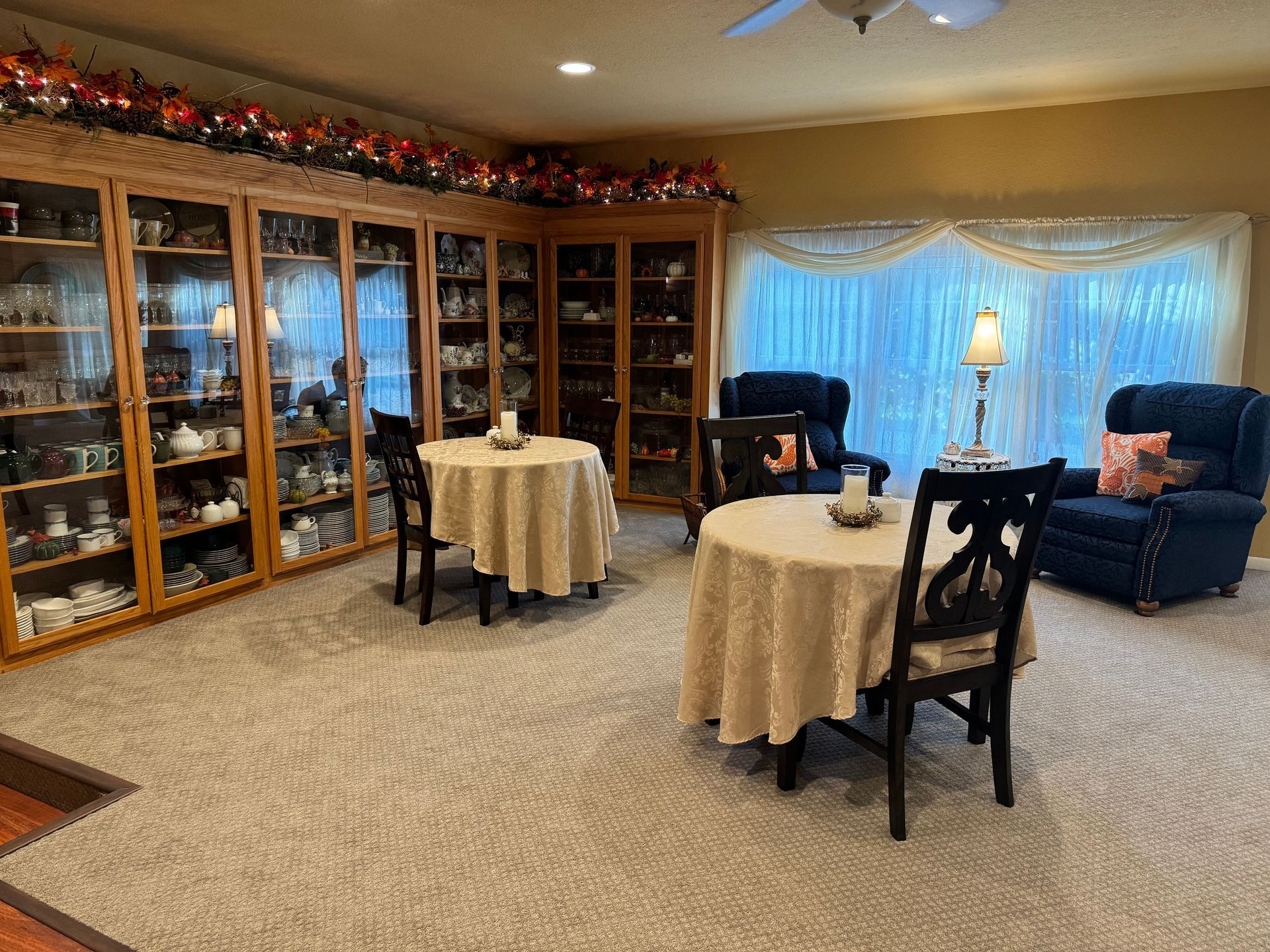Two tables with beige tablecloths, chairs, and two dark armchairs in a room with a large wooden display cabinet.