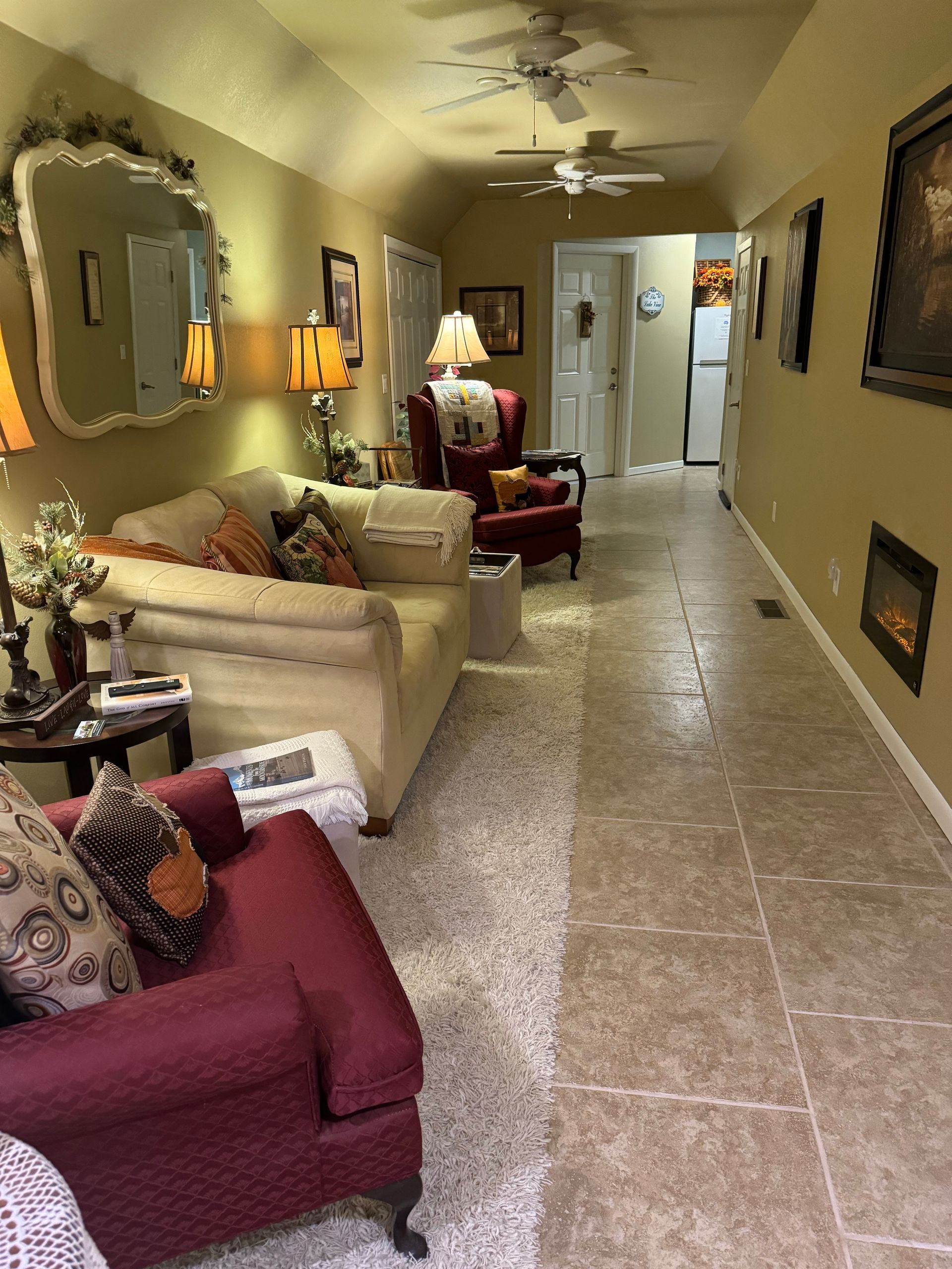 A long, narrow room with a cream sofa, a red armchair, tile flooring, two ceiling fans, and wall art on a beige wall.