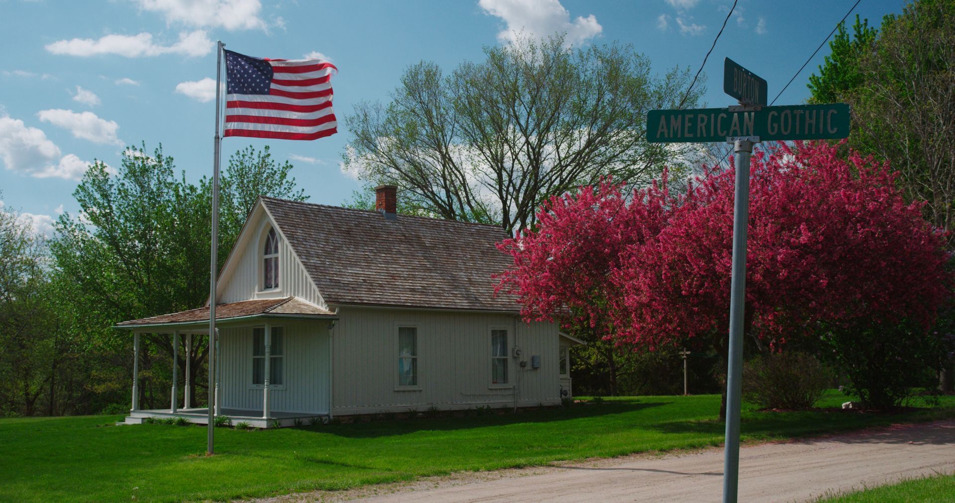 A white house with a porch and an American flag stands near a road sign, with a flowering pink tree on a sunny day.