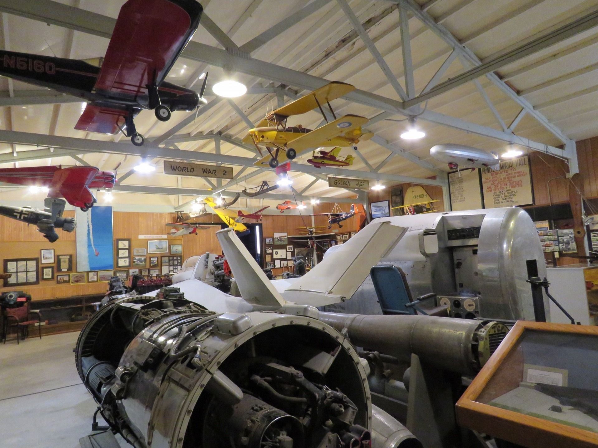 Airplane museum interior with aircraft suspended from ceiling, engine on display.