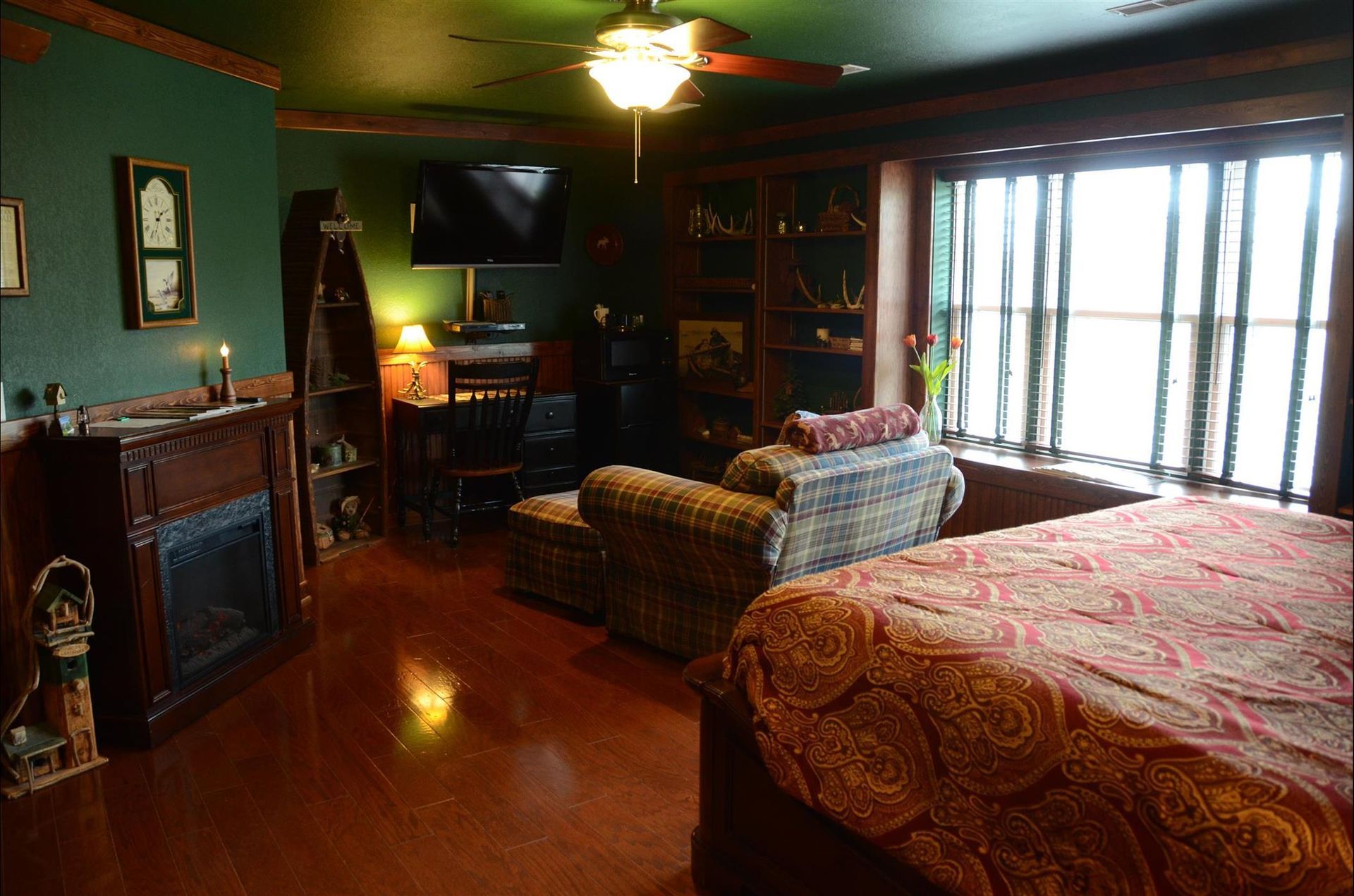 A cozy, dimly lit bedroom with dark green walls, a fireplace, a patterned armchair, and a large bed with a red quilt.
