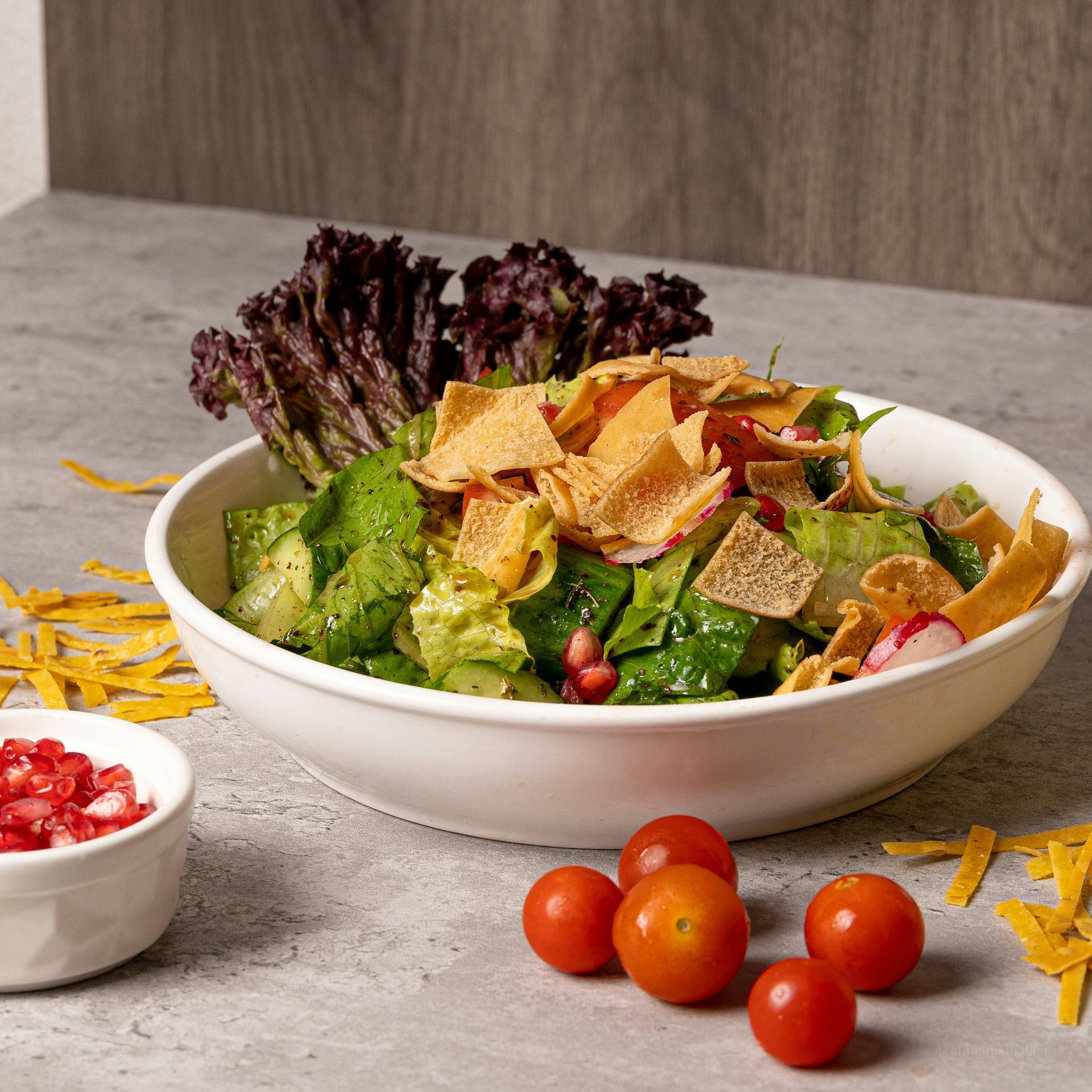 A bowl of fattoush salad with pita chips and pomegranate, served with cherry tomatoes on a gray stone surface.