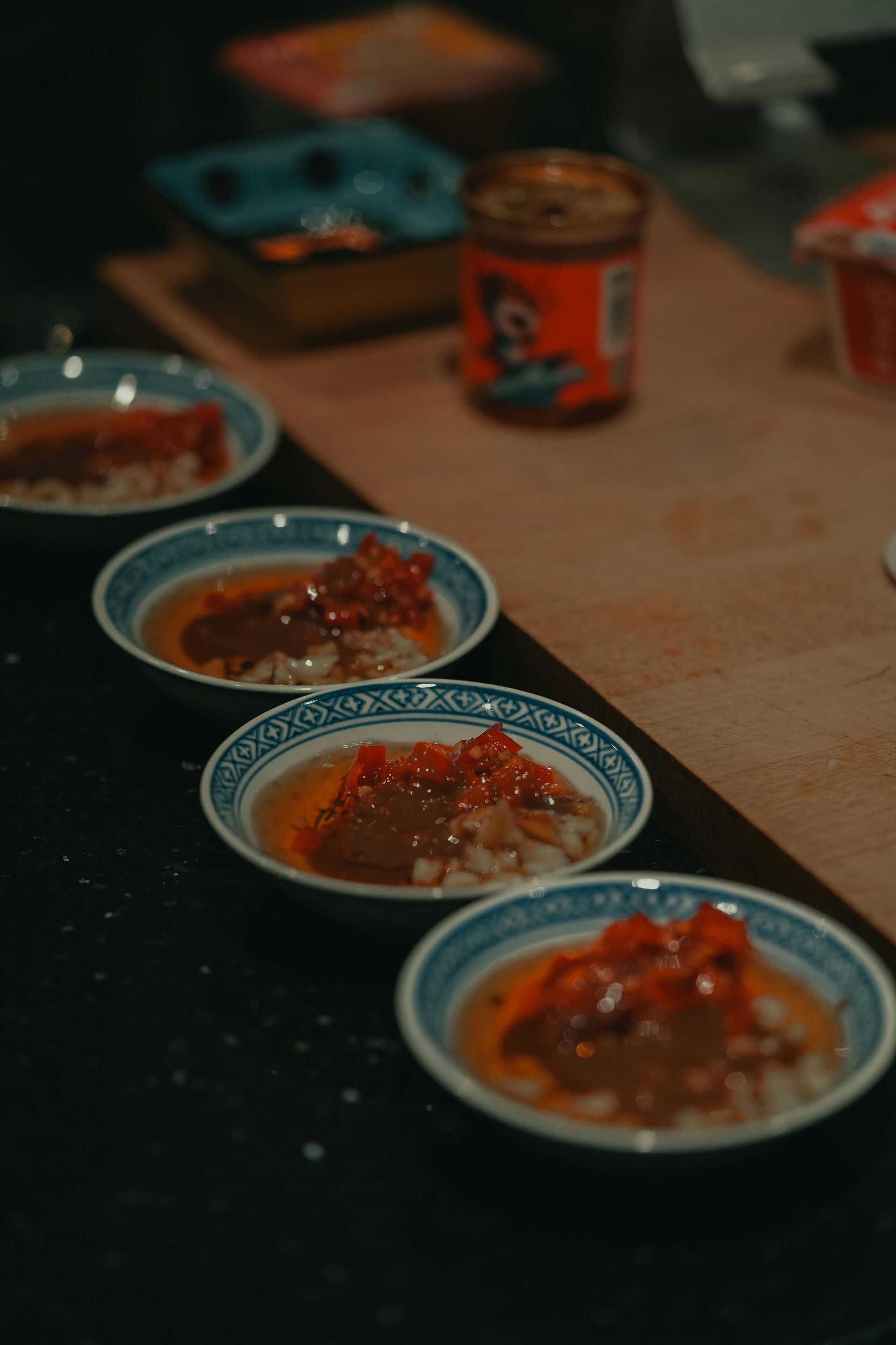 Four small, patterned bowls containing a red sauce and chopped ingredients arranged in a row on a black surface.