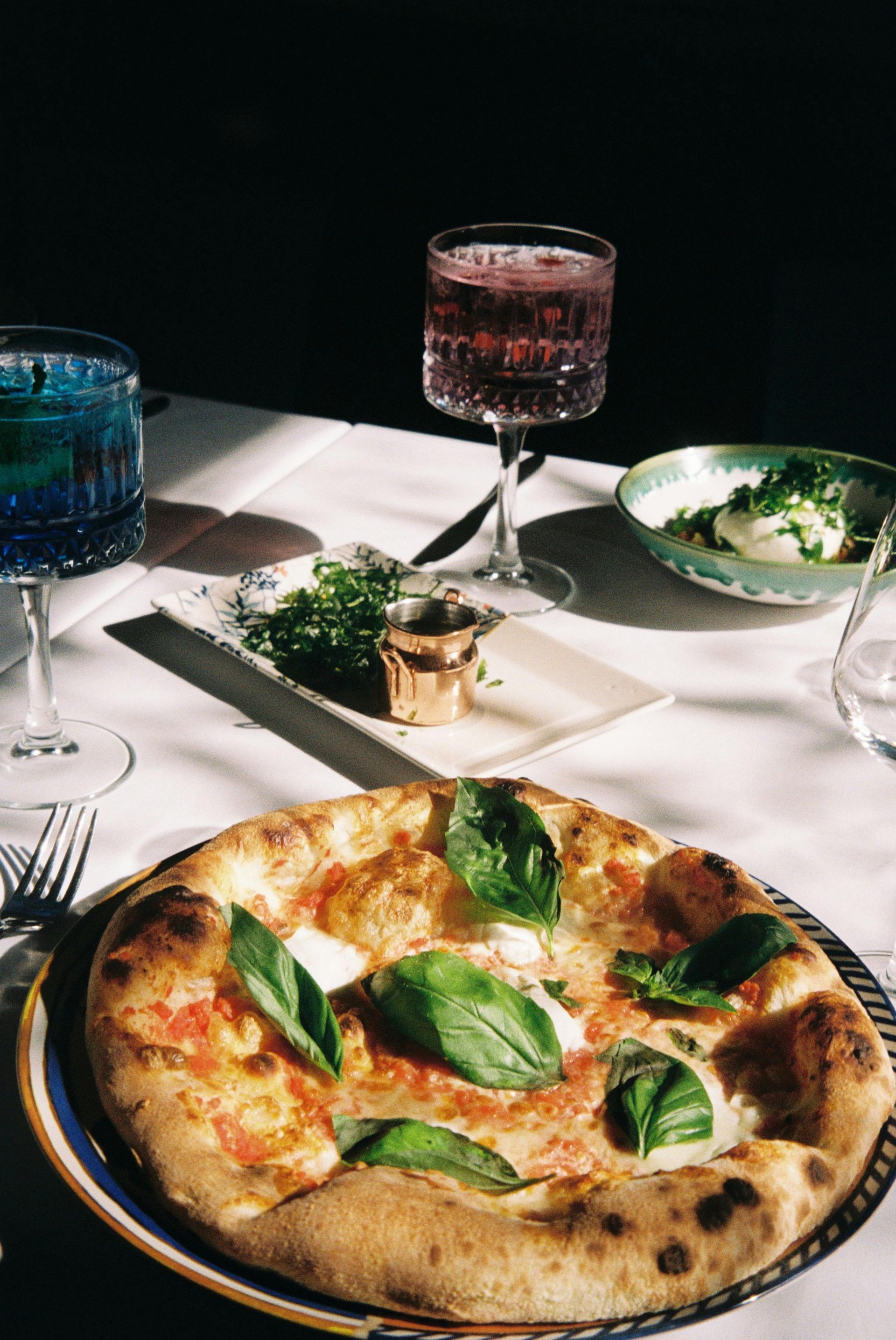 A pizza topped with fresh basil sits on a table next to two cocktail glasses and side dishes in a sunlit setting.