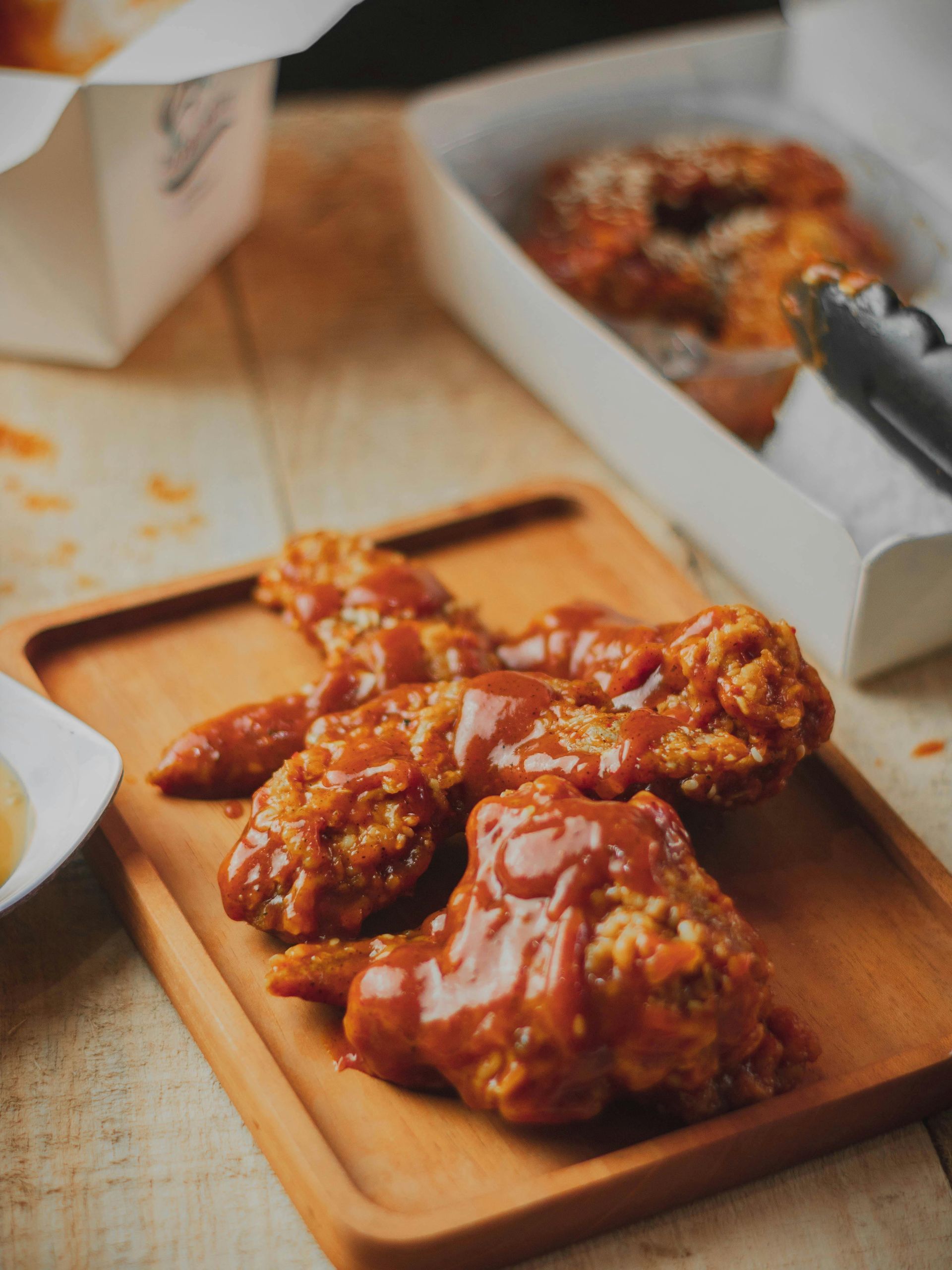 Fried chicken wings coated in red sauce on a wooden board, with takeout containers in the background.
