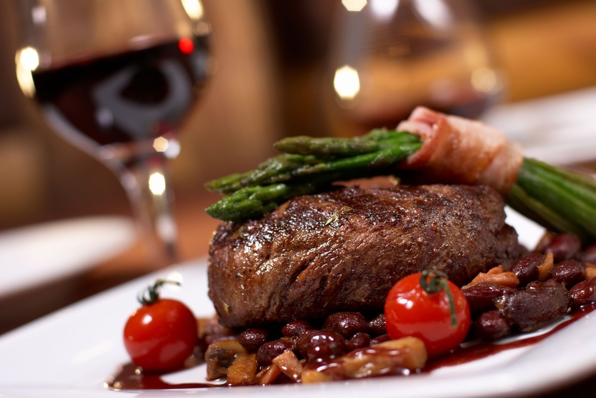 A seared steak topped with bacon-wrapped asparagus and cherry tomatoes on a bed of beans and mushrooms, with red wine.