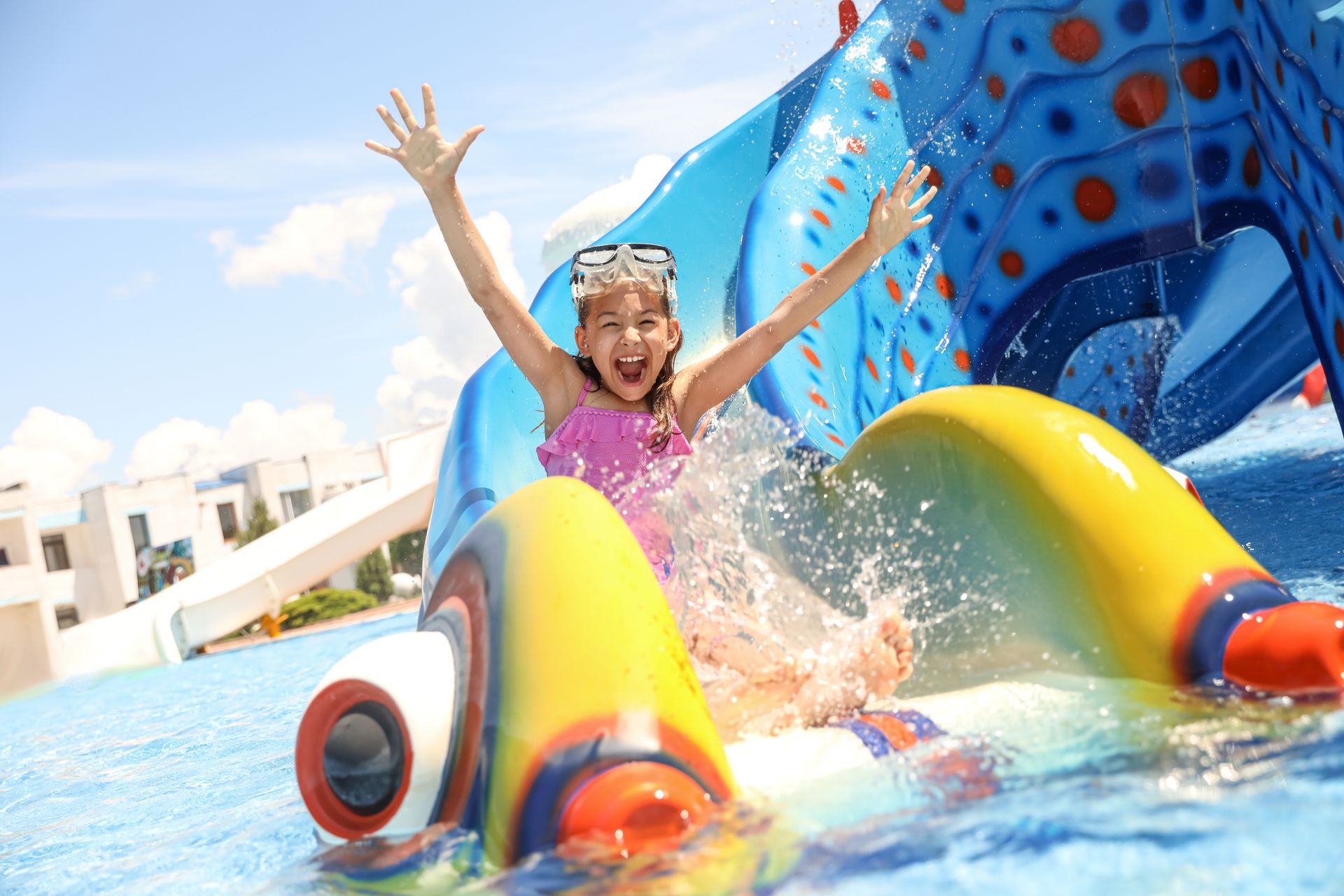 A child in a pink swimsuit joyfully slides down a colorful, fish-shaped water slide at a pool.