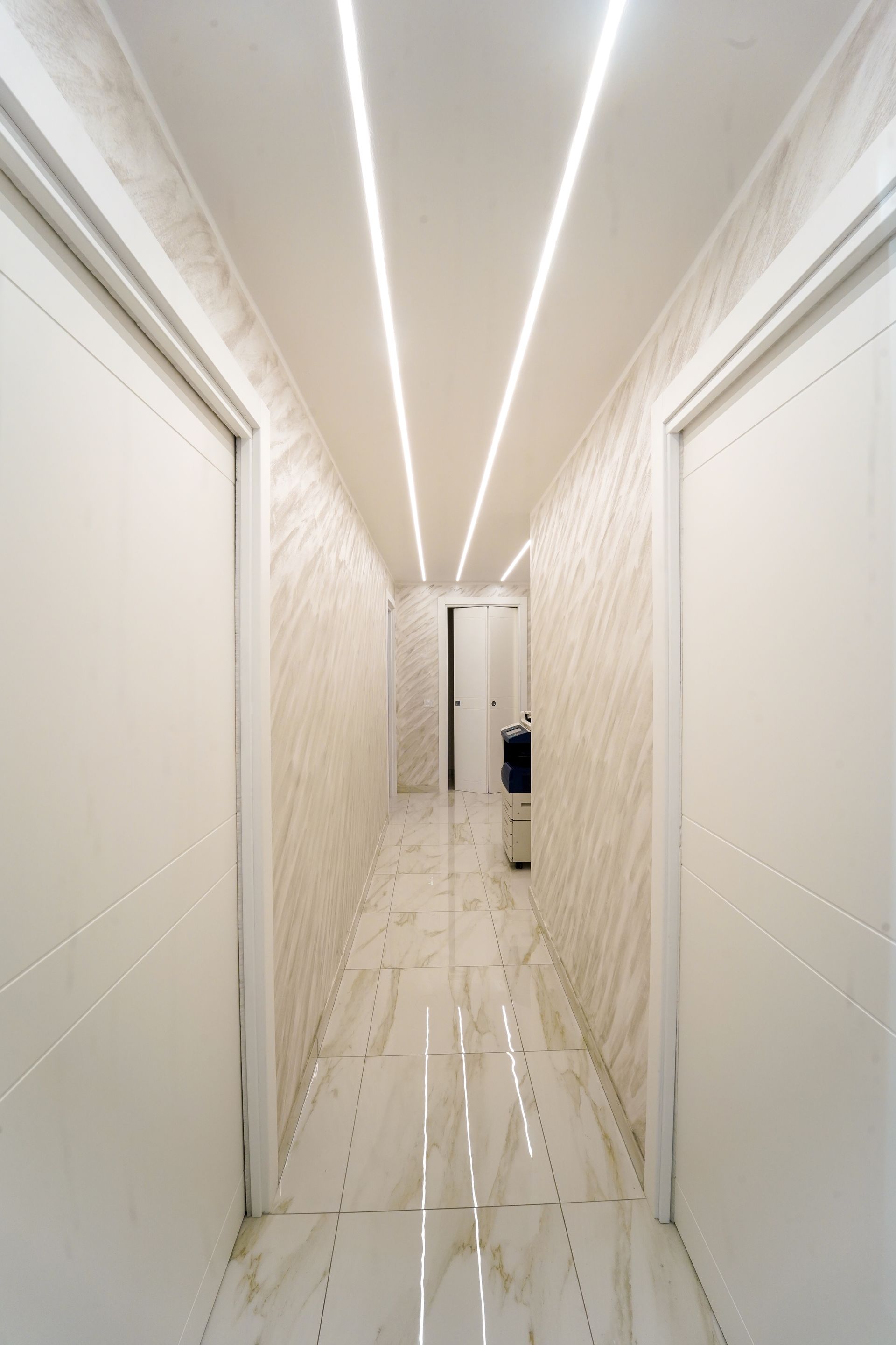 Corridoio lungo e stretto con porte bianche, pareti decorate e luci lineari dall'alto.