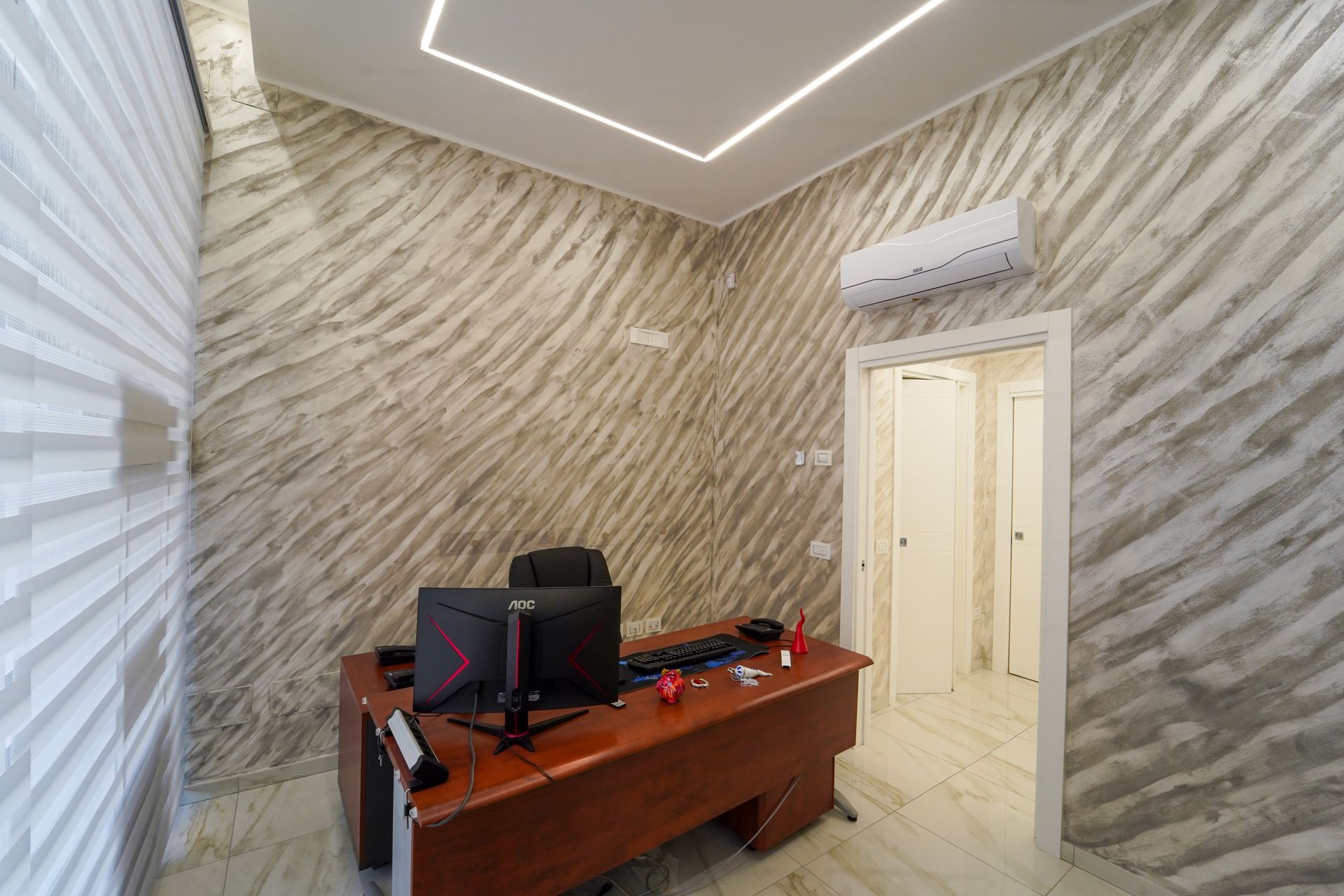 Ufficio con pareti con texture diagonali, scrivania, computer, aria condizionata e porta aperta.