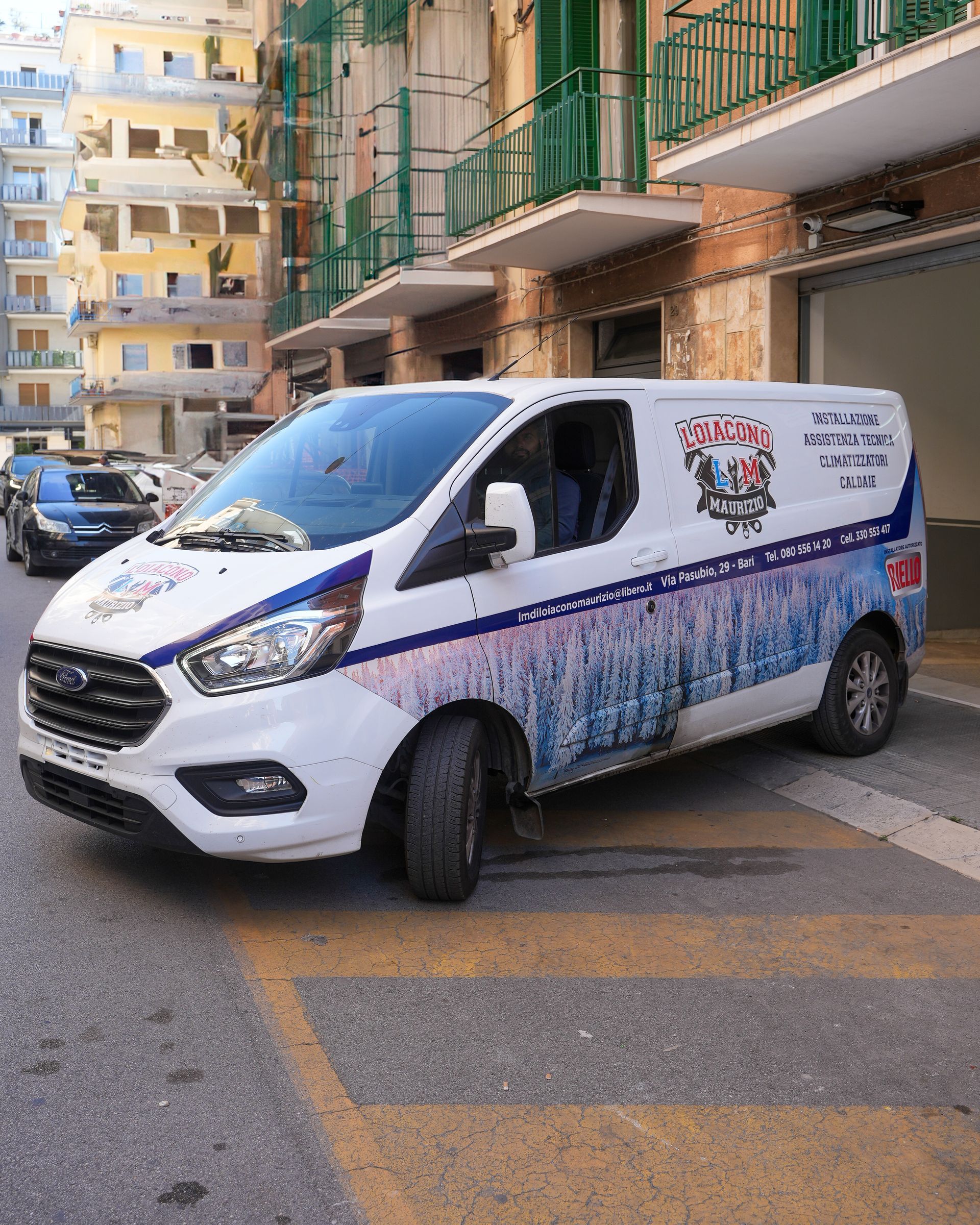 Furgone bianco parcheggiato su una strada con un edificio sullo sfondo; lato decorato con grafica e logo.