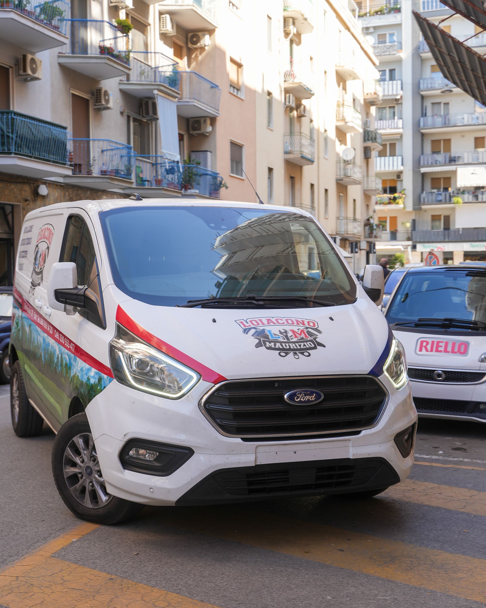 Furgone Ford bianco con logo rosso e blu parcheggiato in strada, con edifici sullo sfondo.