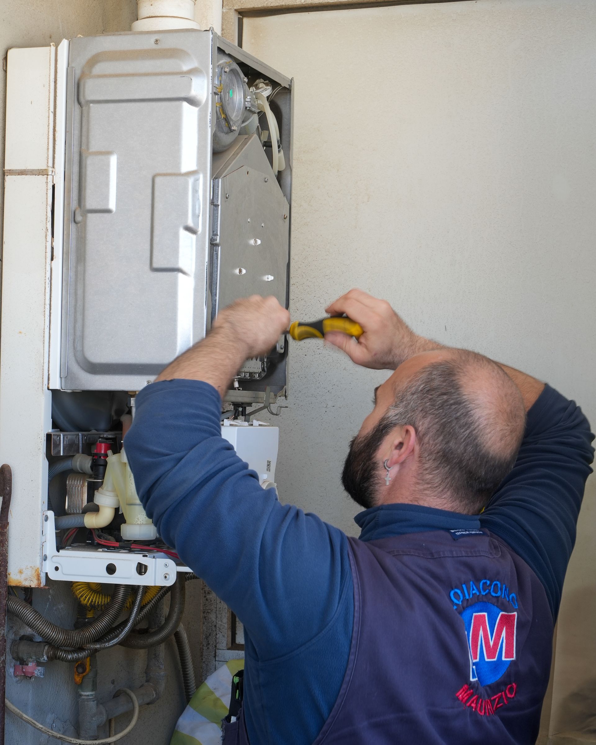 Un idraulico ripara una caldaia a gas montata a parete, utilizzando delle pinze dal manico giallo in una stanza dalle pareti bianche.