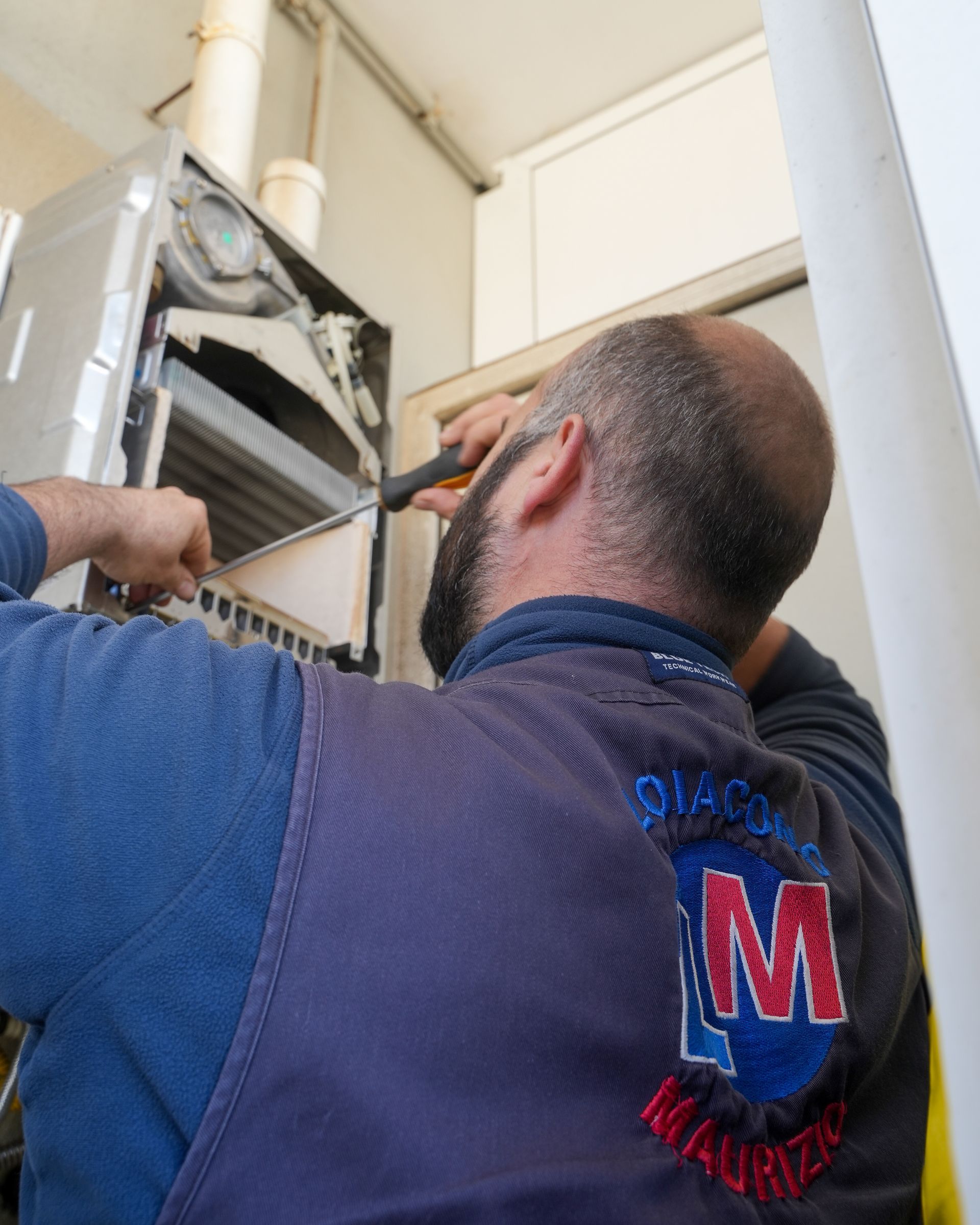 Un uomo con il gilet blu ripara una caldaia a gas con degli attrezzi. Ambiente interno con parete bianca.