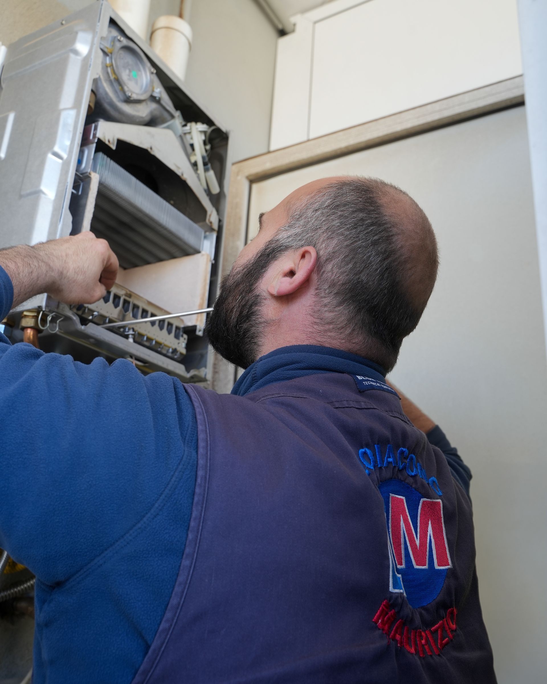 Idraulico che ispeziona una caldaia. Indossa una camicia e un gilet blu e lavora in un ripostiglio.