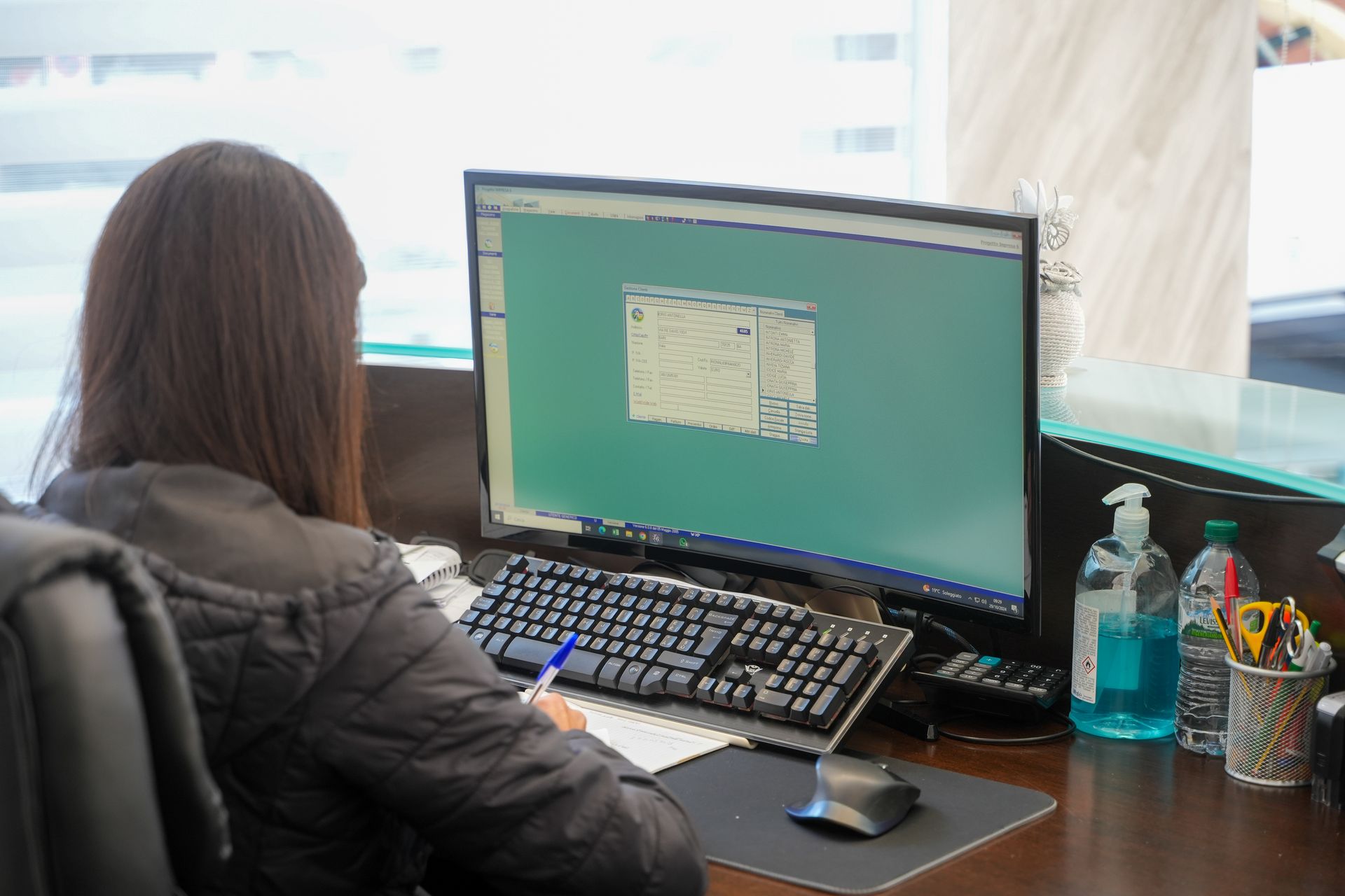 Donna seduta alla scrivania, al computer, che scrive su un foglio. Disinfettante per le mani e penne visibili.