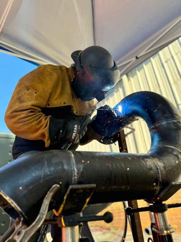 Welder wearing protective gear welding a curved black pipe outdoors. Structural welding services