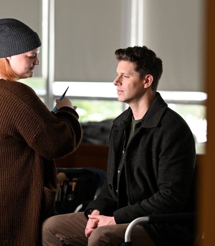 A woman is applying makeup to a man sitting in a chair