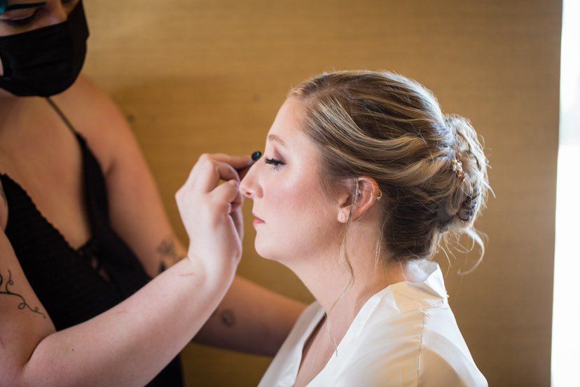 A bride is getting her makeup done by a makeup artist while wearing a mask.