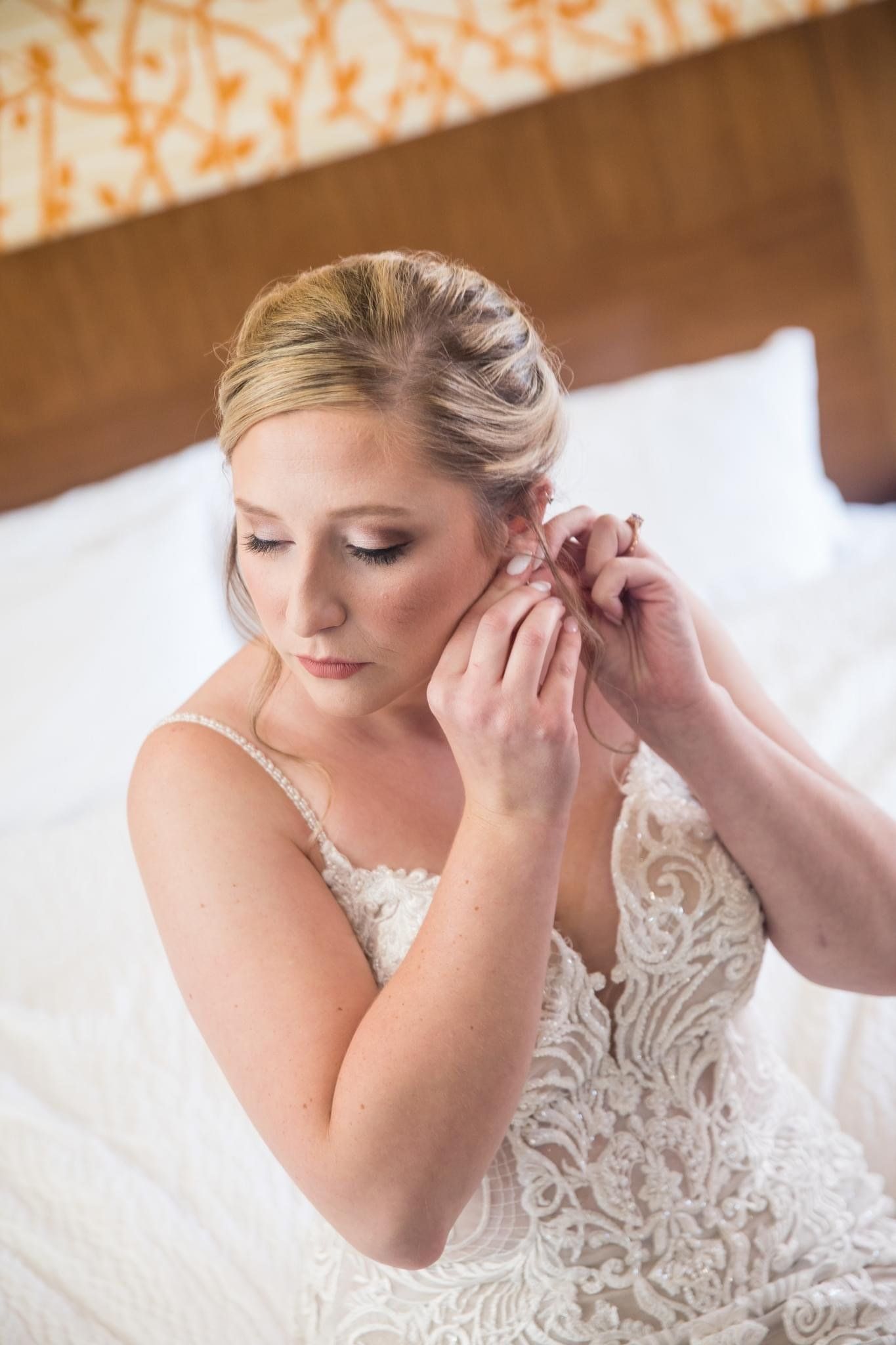 A bride is putting on her earrings while sitting on a bed.