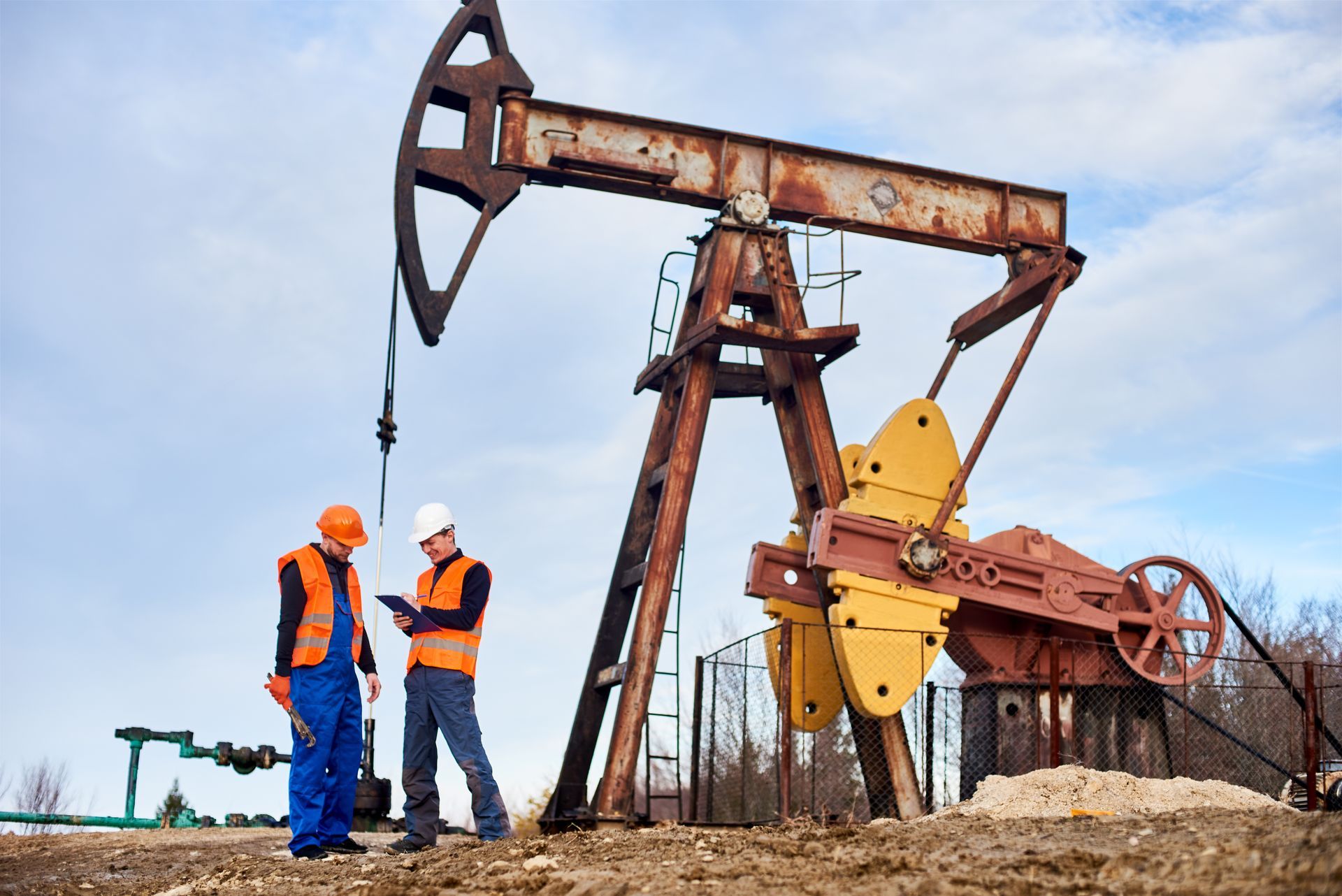 Two workers reviewing documents near a rusty oil pump jack in a field