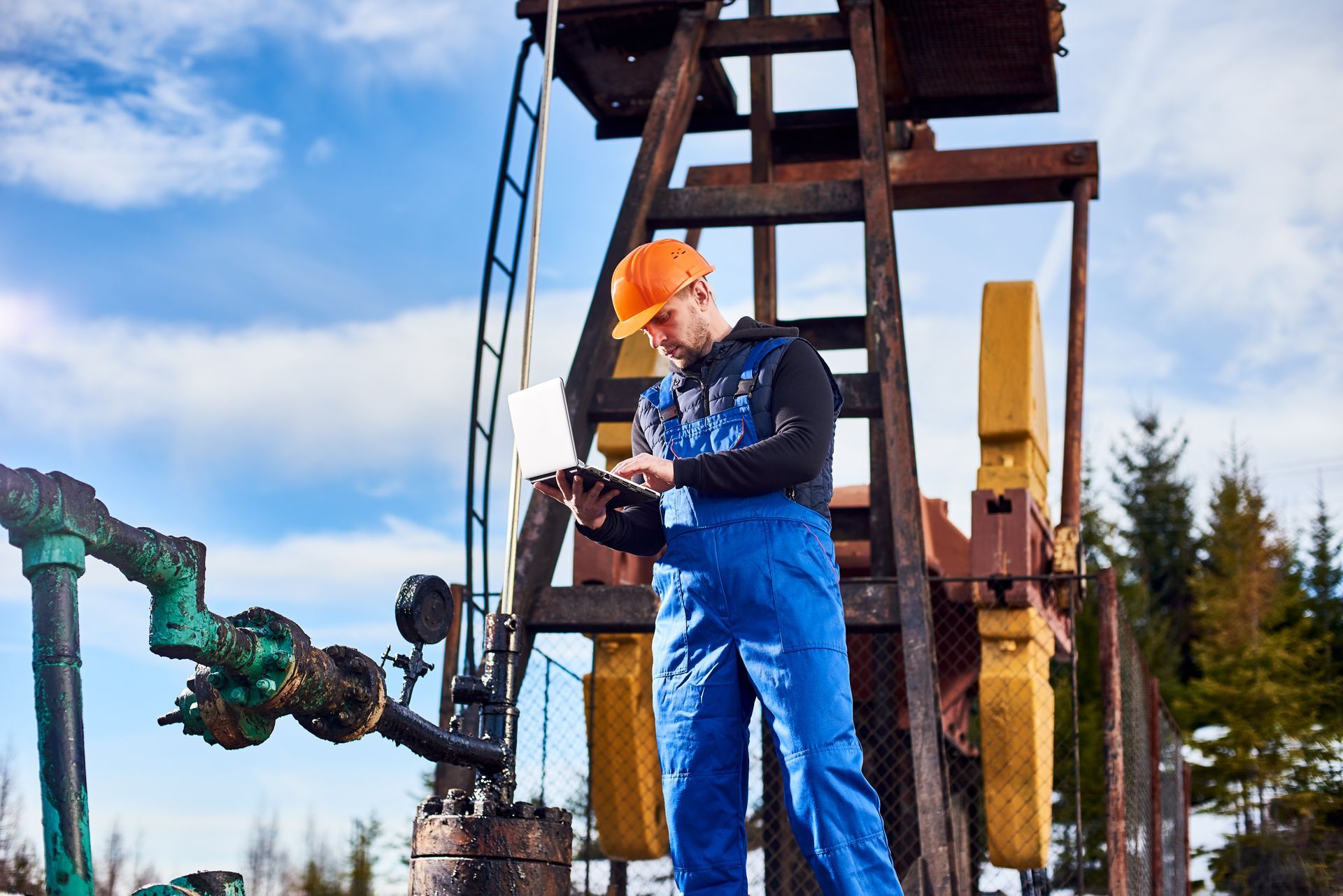 Engineer in blue jumpsuit and orange hardhat uses laptop at an oil rig