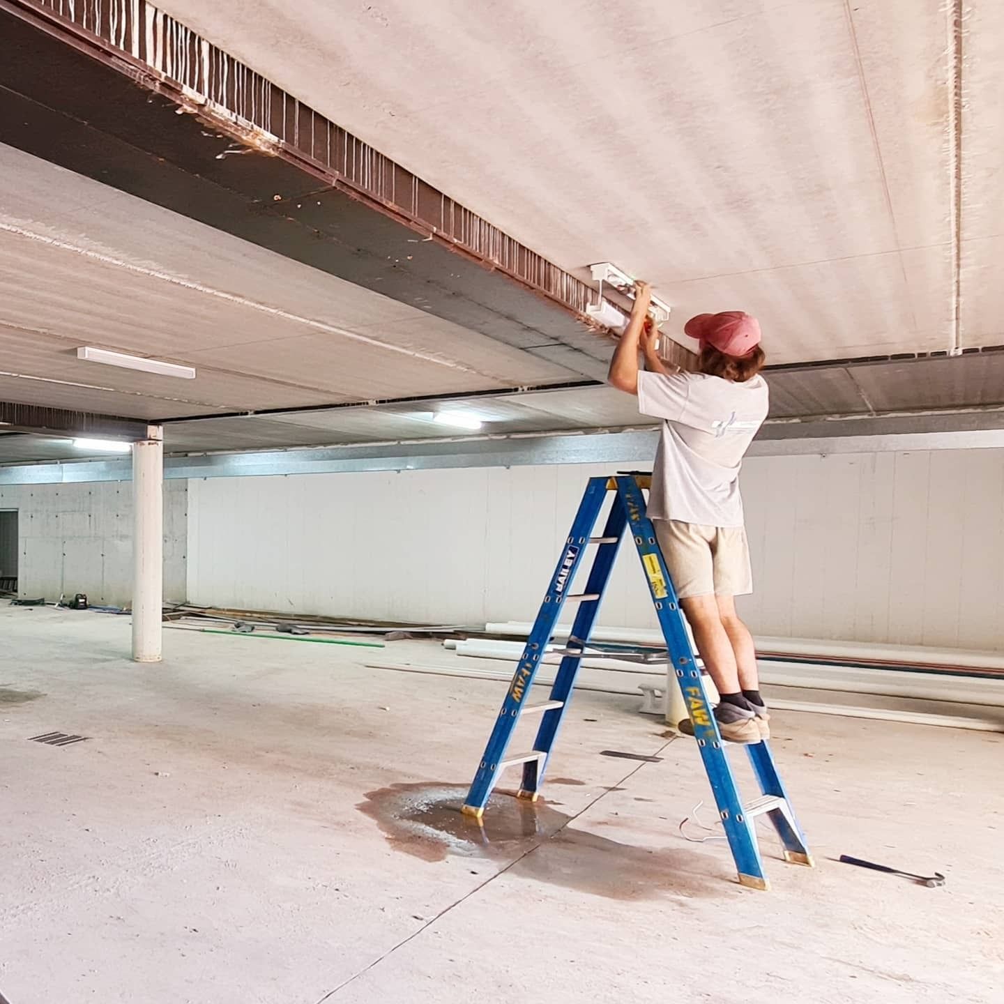 Person on a Blue Ladder Installing a Light Fixture in a Concrete Room — FAW Electrical in Nowra Hill, NSW
