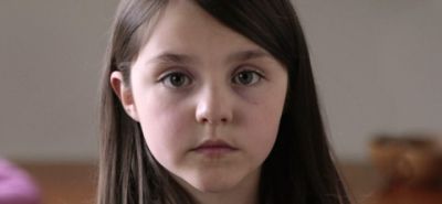 A young girl with long, dark hair stares directly at the viewer; she has bruising under her eyes.