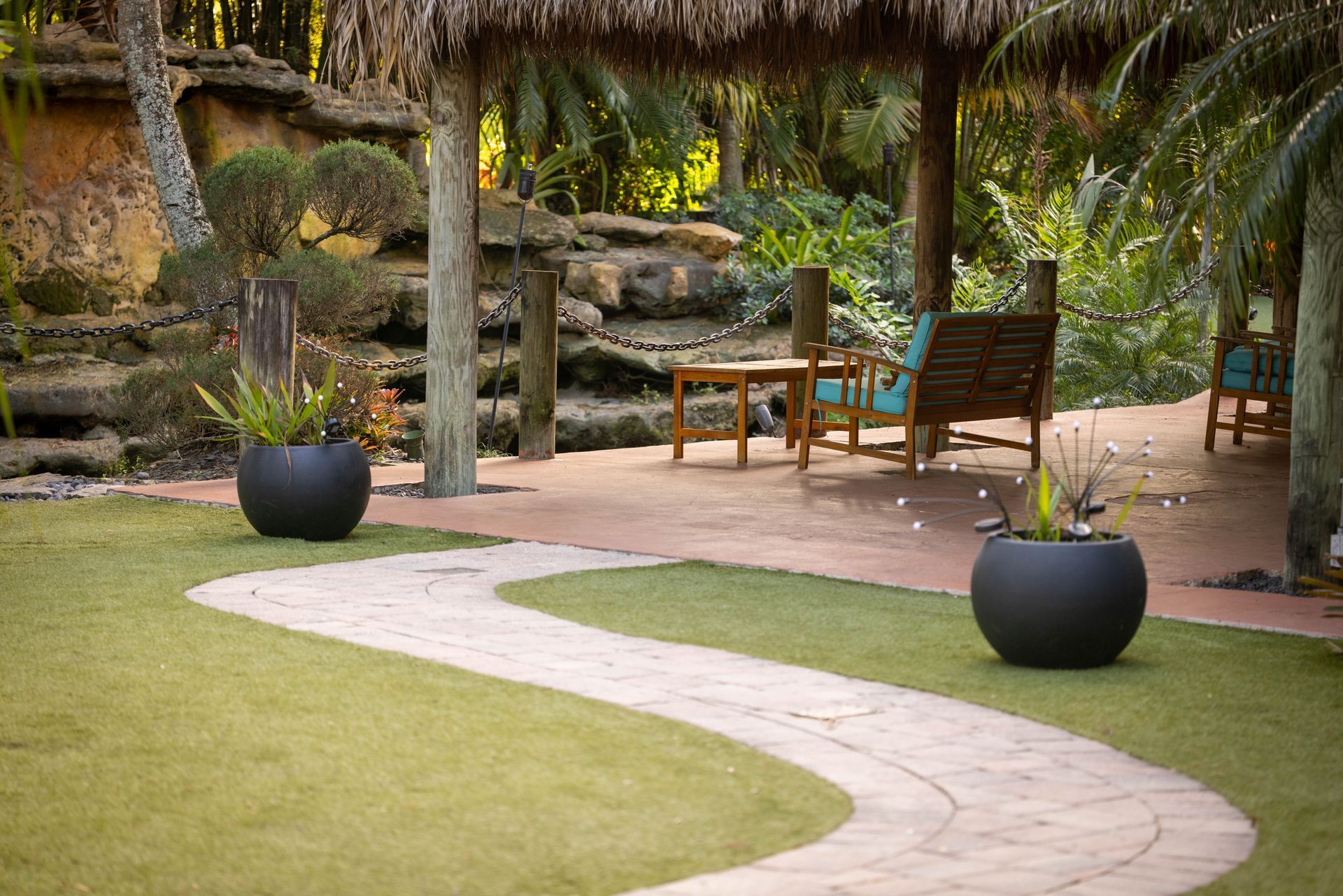 A winding stone path leads to a shaded patio with wooden chairs and planters in a lush, landscaped garden setting.