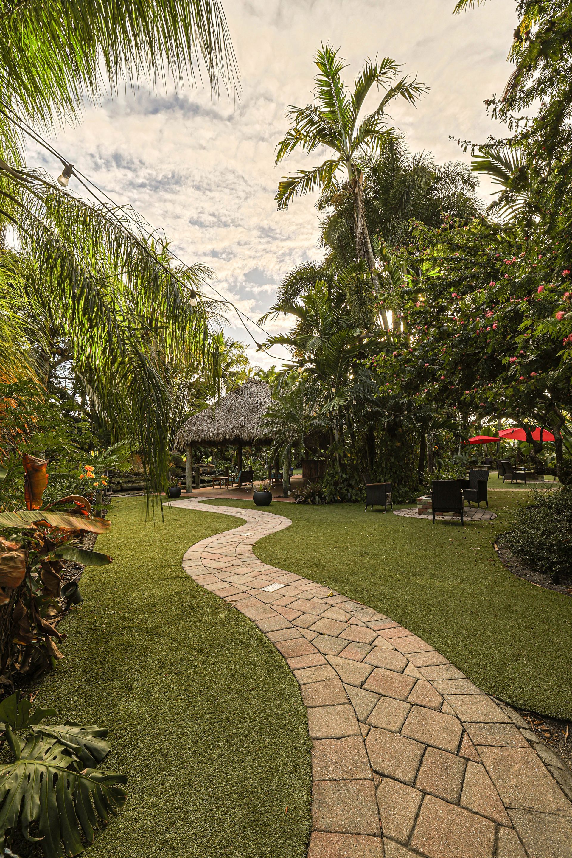 A stone path curves through a lush, green tropical garden toward a thatched-roof gazebo under a cloudy sky.