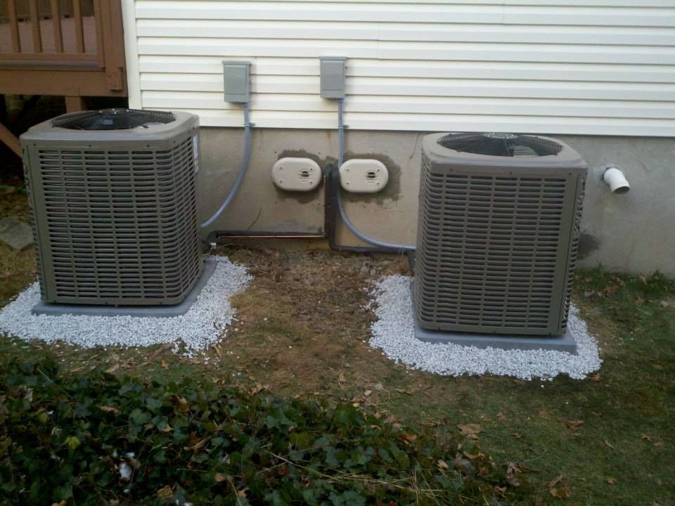 Two grey air conditioning units on gravel pads next to a house with electrical boxes.