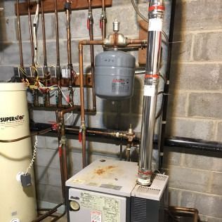 Boiler and water heater in a basement with copper pipes and a silver exhaust vent.