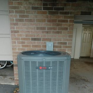 Gray air conditioning unit against a brick wall, electrical box above.