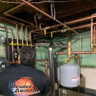 A person in a black shirt examines copper pipes and plumbing. A water tank and valves are also visible.