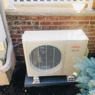 Fujitsu heat pump unit next to a brick wall and gutter. Beige unit sits on concrete blocks.